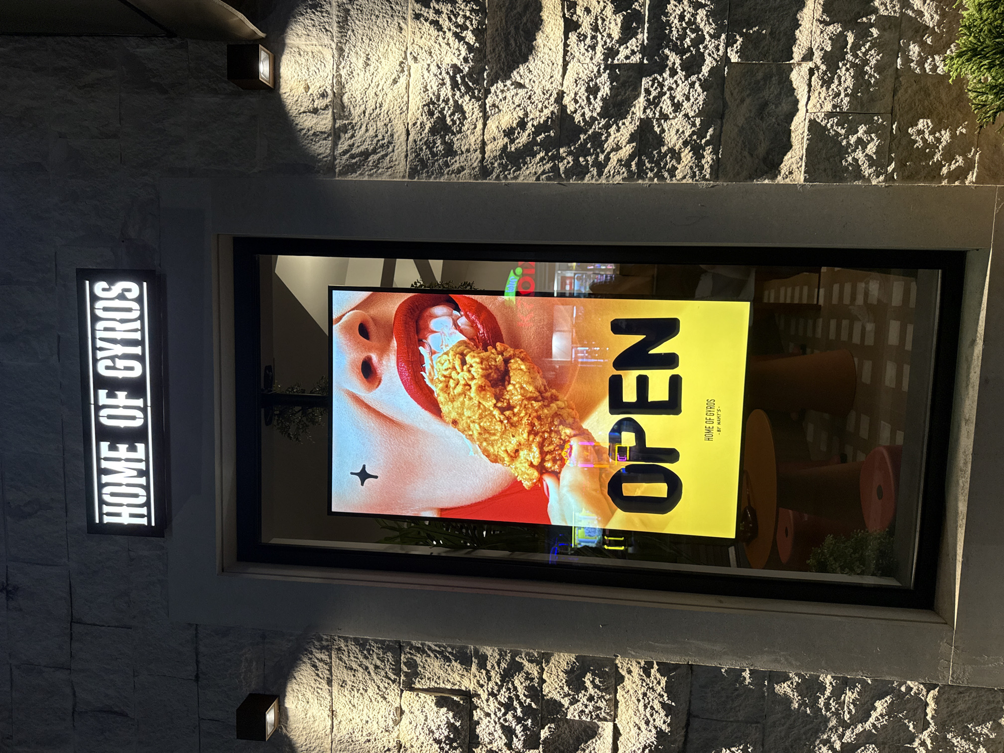 The image shows the exterior of a restaurant named "HOME OF GYROS" in Budva, Montenegro. The restaurant has a modern facade with stone-like cladding. Above the entrance, a rectangular illuminated sign displays the restaurant's name in white lettering against a black background. The main focal point is a large digital screen or window display within the entrance. This display shows a close-up of a person's mouth with bright red lipstick biting into a piece of fried chicken. Below this image, the word "OPEN" is prominently displayed in large, bold yellow letters. Beneath "OPEN," smaller text reads "HOME OF GYROS" and "- BY NAKI'S -". Two outdoor spotlights illuminate the facade. Through the glass, interior elements like orange tables and stools, as well as some greenery, are visible. The overall scene suggests the restaurant is open for business at night.