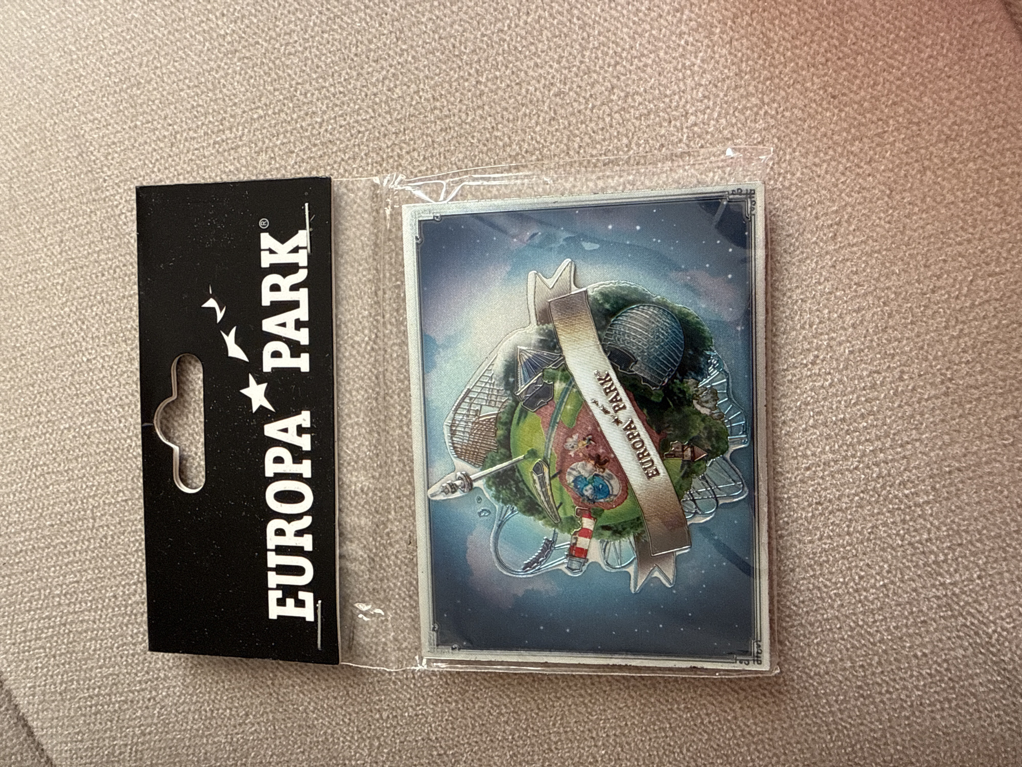 The image displays a new, packaged souvenir item from Europa-Park, laid flat on a light beige, textured surface, possibly a carpet or fabric.
The main subject is a rectangular, clear plastic package containing a unique decorative item. To its left, a black cardboard backing is partially visible, featuring the prominent "EUROPA PARK" logo. The logo consists of "EUROPA" stacked above "PARK", with a stylized star replacing the 'O' in Europa and two small bird-like shapes to the left of "EUROPA". A registered trademark symbol (®) is visible next to "PARK". The cardboard also has a typical retail hanging hole cut-out.
The decorative item inside the plastic package appears to be a magnet or pin. It depicts a miniature, spherical world, intricately designed with various elements characteristic of an amusement park. Visible details on this miniature world include roller coaster tracks, a large geodesic dome structure, a lighthouse, and other small buildings and trees, all integrated into a green, lush landscape. A metallic-looking banner stretches diagonally across this miniature world, prominently displaying "EUROPA PARK" in white text. The background of the decorative item itself is a dark blue night sky filled with small white stars, creating a whimsical and dreamy aesthetic for the miniature scene.
No activity is taking place; the image is a static product shot. The lighting is bright and even, highlighting the details of the item. The item appears to be in pristine condition, still sealed in its original packaging. The close-up crops provide clearer views of the "EUROPA PARK" text on both the packaging and the souvenir itself, as well as the texture of the background surface and the edges of the plastic wrapping.