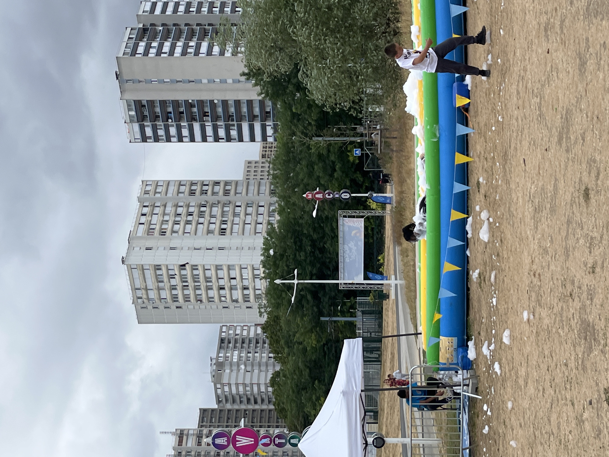 The image depicts an outdoor scene with several apartment buildings in the background, characteristic of the urban landscape of Bobigny, France. In the foreground, an inflatable pool filled with foam is set up. A young boy, wearing a white t-shirt and dark pants, is walking near the pool. Several other people are visible in the middle ground, including children near a white tent structure and individuals standing by a fence. A signpost with circular emblems is also visible on the left side of the image. The ground is covered in sand or dirt, with scattered white foam pieces. The sky is overcast.