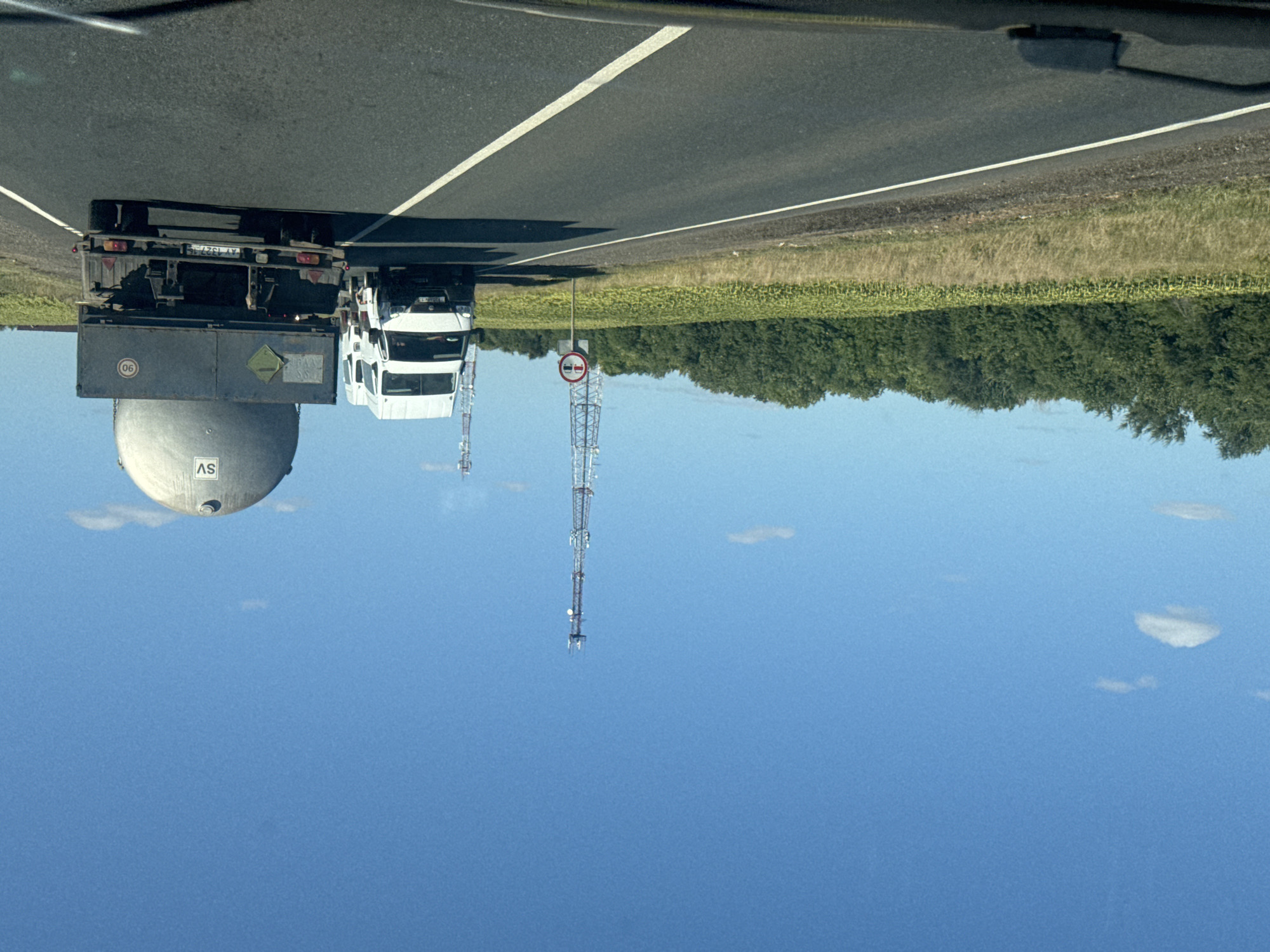 A clear, blue sky with scattered clouds stretches over a rural landscape. In the foreground, a two-lane asphalt road is visible, with white lane markings. On the right side of the road, a large truck is transporting a spherical tank. To the left of the truck, a white van is stopped, and further down the road, another white van is visible. A field of sunflowers lines the right side of the road, with a dense line of trees beyond it. Two telecommunication towers rise from the background. A traffic sign indicating "no overtaking" is positioned on the left side of the road. The image appears to have been taken from a moving vehicle. The location is identified as Perevolotsky, Russia.