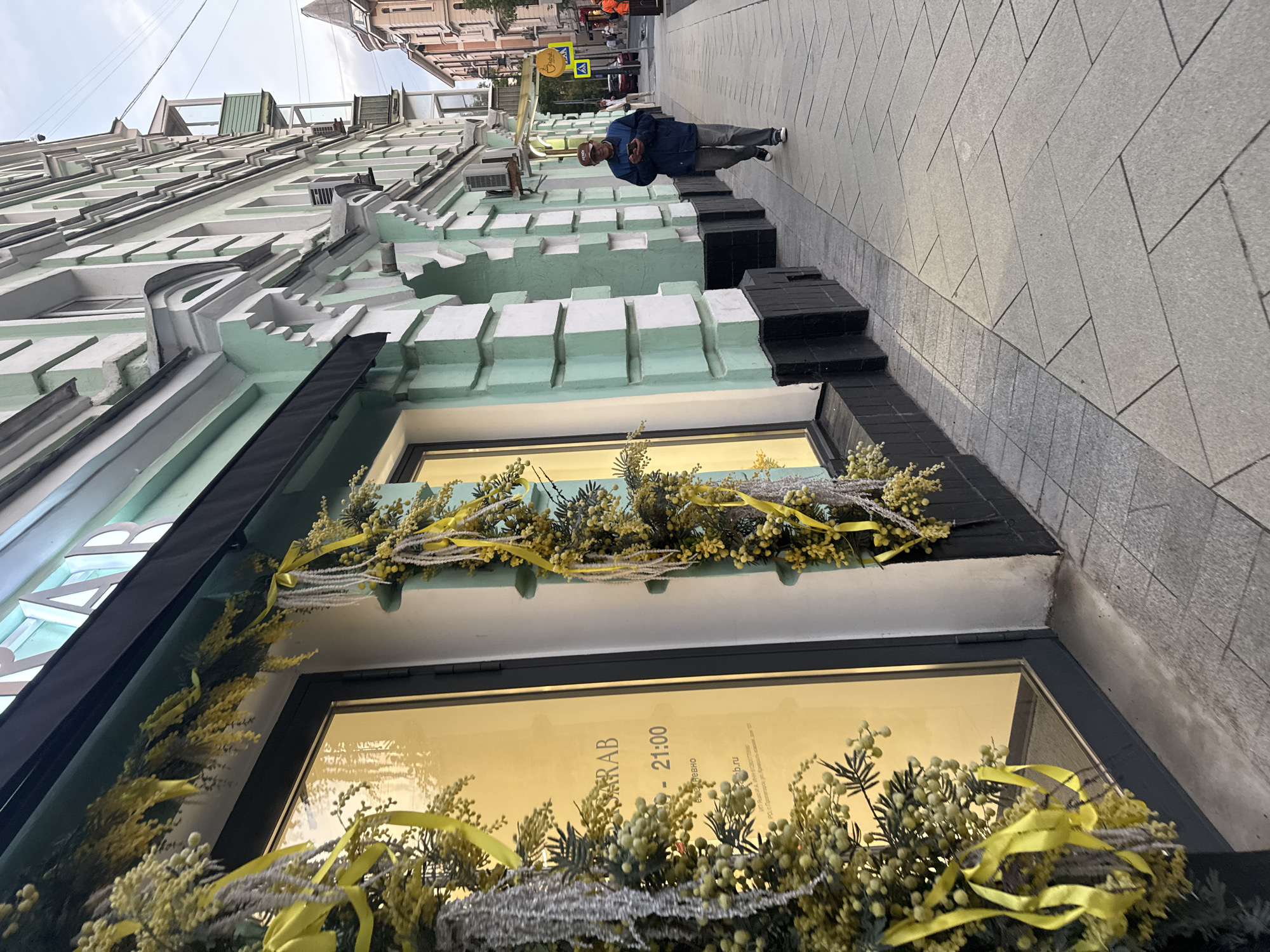 A man walks down a paved sidewalk in Moscow, Russia, outside a building with a light green and white facade. The building has large windows decorated with garlands of yellow flowers and ribbons. The man, wearing a blue jacket, gray pants, and a baseball cap, looks at his phone as he walks. The sky is overcast.