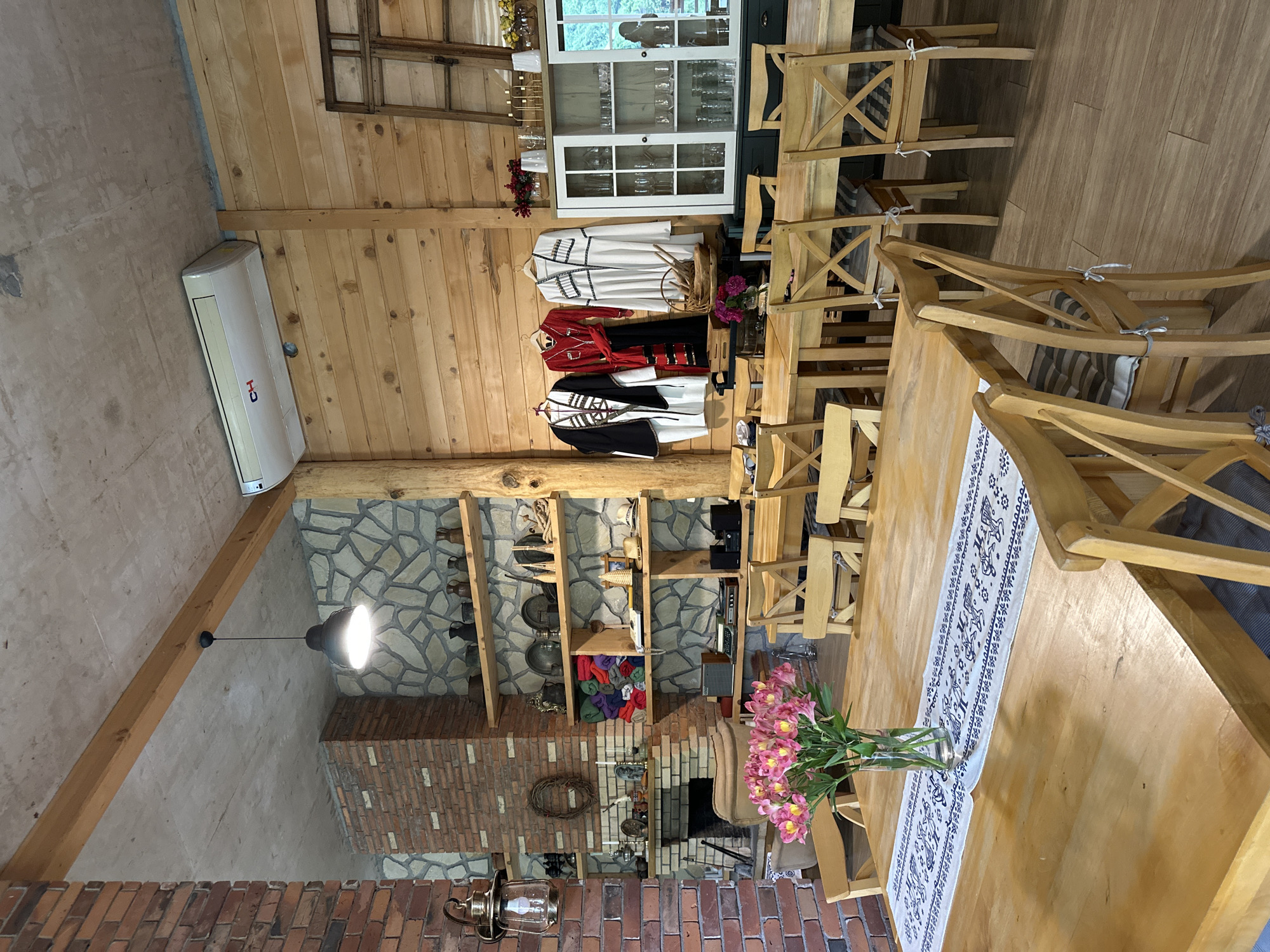 The image captures the interior of a room, likely in Tibeta, Georgia, evident from the traditional clothing displayed. The room features a wooden-paneled wall, interspersed with a stone section that includes a brick fireplace and open shelving. A collection of objects and souvenirs adorn the shelves. Traditional Georgian costumes are hanging on the wall. The room has a large wooden table, set with a white runner. A vase of pink flowers sits in the center of the table. Wooden chairs surround the table. An air conditioning unit is mounted high on the wall.