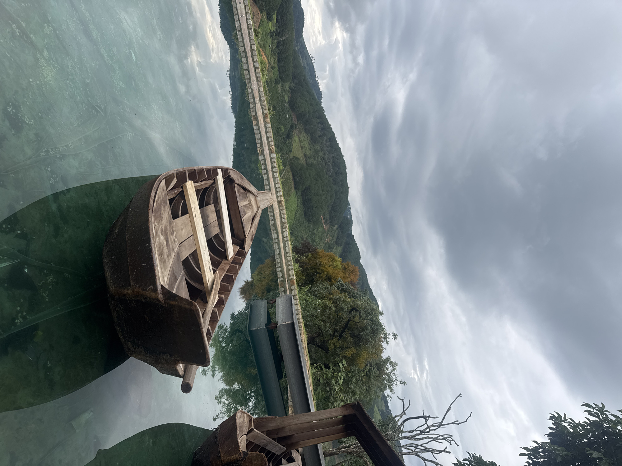 The image captures a serene, overcast day with a cloudy sky. In the foreground, two wooden boats are gently floating on calm, clear water, their reflections shimmering on the surface. One boat, positioned closer to the viewer, is empty and shows its interior with wooden slats. The other boat, partially visible on the right side of the frame, also appears empty.
The water is bordered by a stone wall, beyond which a lush green hillside rises. The hillside is covered with dense trees and vegetation, with evidence of terraced farming or cultivation. The landscape extends into the distance, with rolling hills under the muted light of the cloudy sky.
To the right, a rustic wooden structure, possibly a pier or part of a building, is visible, adding a man-made element to the natural scene. A bare, gnarled tree branch extends into the sky from this side, contrasting with the vibrant green of the surrounding foliage.
The overall impression is one of tranquility and natural beauty, with the boats suggesting a possibility for leisure or transport across the water, set against a backdrop of verdant hills. The location is identified as Thôn 3, Vietnam, a place likely characterized by such natural landscapes and waterways.