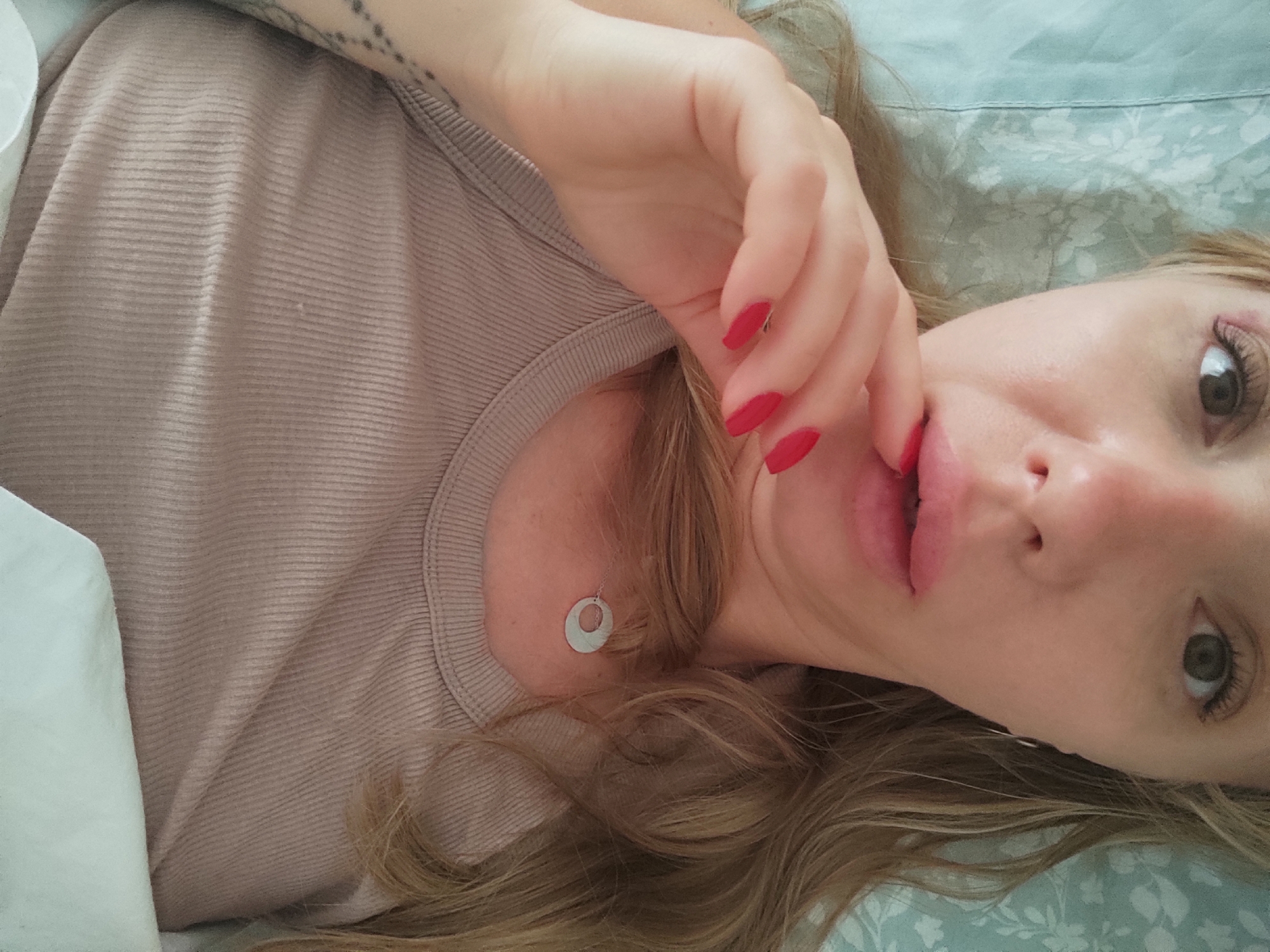 The media file is a close-up, slightly angled, over-the-shoulder selfie of a young woman lying down. She has blonde hair and appears to be in a relaxed or playful pose, with her right hand near her mouth and her fingers painted with red nail polish. Her eyes are open and looking towards the camera. She is wearing a light beige, ribbed tank top and a delicate circular pendant necklace. The background suggests a bed with patterned light blue and white bedding. The lighting is soft, indicating it might be daytime. There is no visible text in the image. The location context provided is Blanes, Spain, but there are no specific visual cues within the image to confirm this.