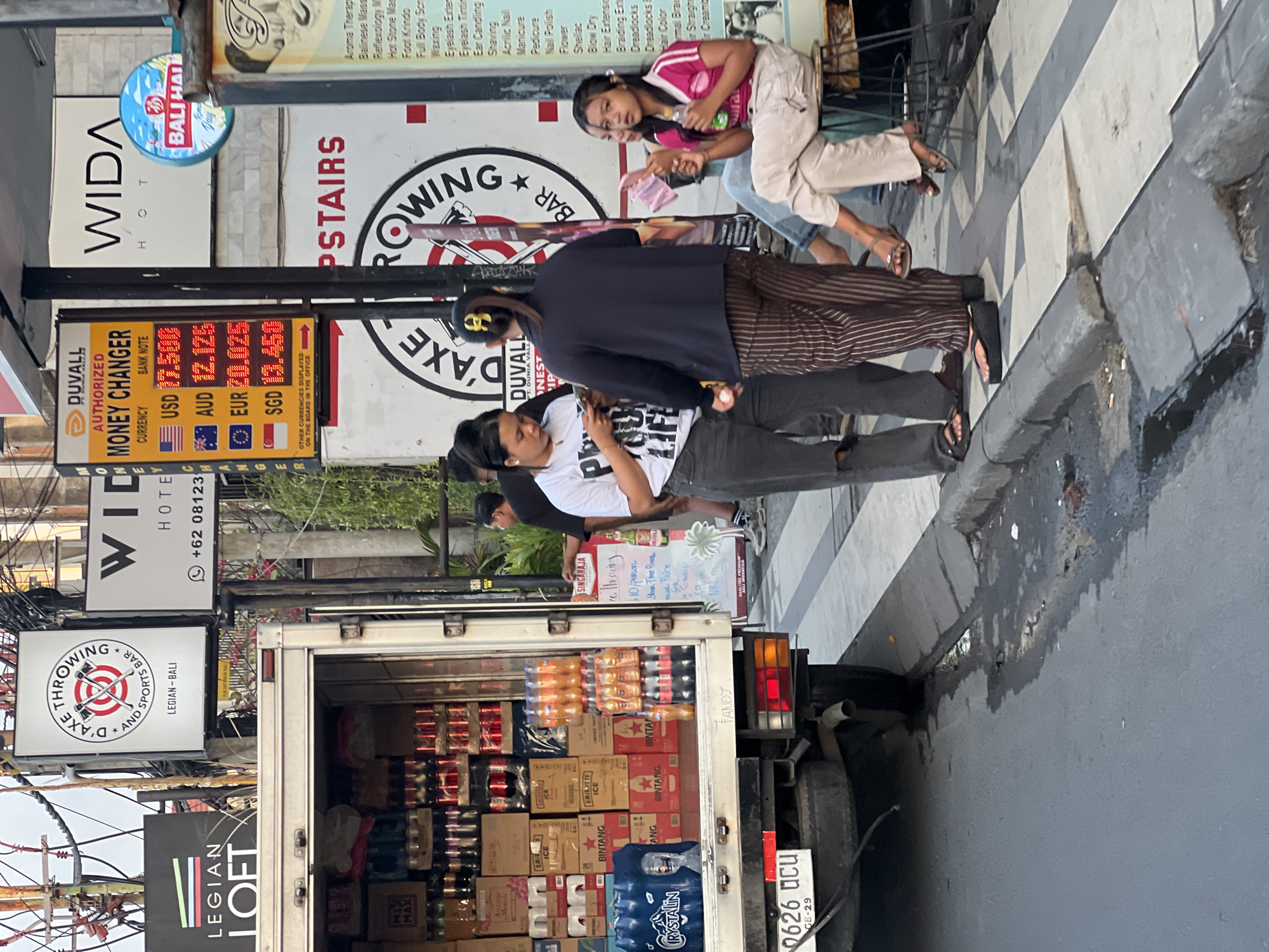 The image depicts a street scene in Kuta, Indonesia, likely during the daytime. A delivery truck, partially loaded with beverages and other goods, is parked on the side of the road. Several people are visible in the foreground and background.
In the foreground, a woman in a white t-shirt with text on it and dark pants is looking at her phone. She is wearing dark sandals. Beside her, a woman in a dark top and striped pants is standing and looking away. She is wearing dark platform sandals. Further to the right, two young women are seated on stools. One woman, wearing a pink top and light-colored pants, is holding a bottle and looking towards the left. The other woman is partially visible and is wearing a blue top and light-colored pants.
Above the delivery truck, several signs are visible. One sign reads "LEGION LOFT." Another sign is for "D'AXE THROWING AND SPORTS BAR" with an image of crossed axes. A money changer sign displays exchange rates for USD, AUD, EUR, and SGD. A sign for "WIDA HOTEL" is also present, along with a circular sign with "BALI HAI."
The environment appears to be a busy street with electrical wires and lush greenery visible in the background. The road surface is asphalt, and a sidewalk with a checkered pattern is present. Debris and water are visible on the side of the road. The license plate of the truck reads "0626 UCU."