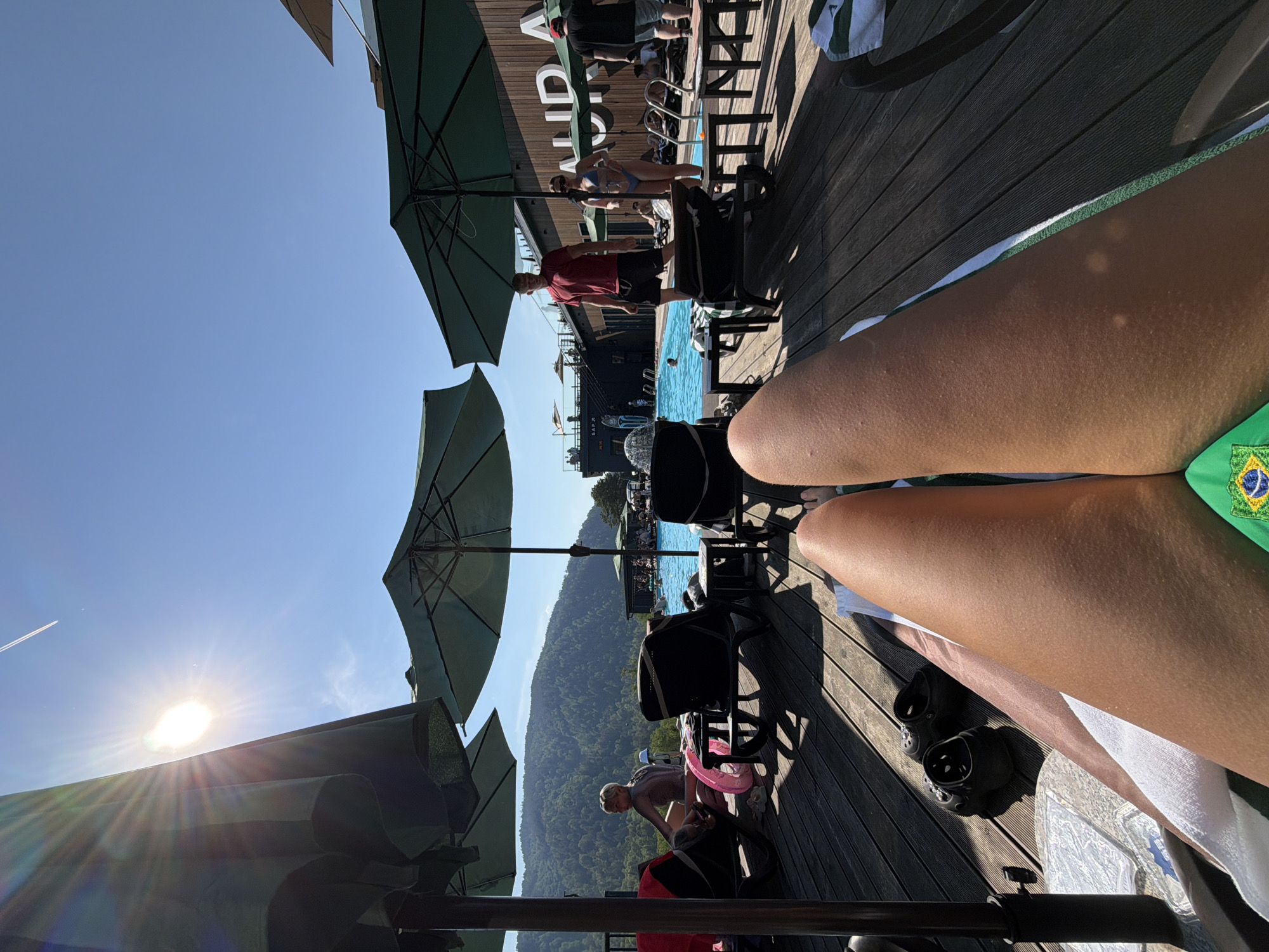 This image depicts an outdoor leisure scene at the Aura Resort Hotel & Spa in Divnogorsk, near Манский in Krasnoyarsk Krai, Russia, under a clear, bright blue sky. The perspective is a first-person view from a person relaxing on a sun lounger.
In the immediate foreground, the person's tanned legs, exhibiting numerous small bumps consistent with goosebumps or piloerection, are prominent. They are wearing green swimwear with a yellow and blue Brazilian flag emblem on the visible portion. To the left of the legs, a pair of black Crocs and a white towel rest on the dark wooden decking that covers the ground.
The mid-ground showcases a large, rectangular blue swimming pool. The pool area is furnished with numerous dark-colored sun loungers and small tables, many positioned beneath large green sun umbrellas providing shade. Several individuals are present in this area. On the far left, a person with short, light-colored hair, wearing a light-toned, long-sleeved swimsuit, sits on a sun lounger beside a pink inflatable ring. In the center-right, a man in a red t-shirt and black shorts stands near the pool's edge, facing left, while a woman in a light blue bikini stands slightly behind him. Further to the right, a man in a black t-shirt and grey shorts is partially visible. Multiple other individuals are visible swimming in the pool and relaxing on loungers around its perimeter.
In the background, a multi-story building with dark wooden cladding and large white letters spelling "AURA" is visible, indicating the resort's name. A darker structure to the left of the main building has partial Cyrillic text, "БАР ?" (BAR ?), suggesting a bar. Behind the resort structures, a vast expanse of densely forested, green mountains dominates the horizon under the bright sky. The sun, positioned in the upper left portion of the image, causes a significant lens flare. A faint white aircraft contrail is also visible in the upper left quadrant of the sky.