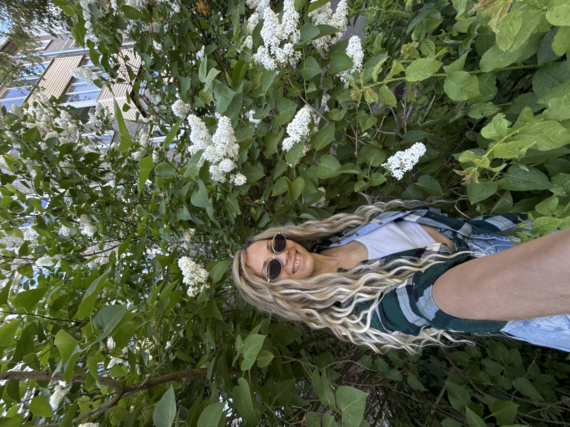 A woman is smiling at the camera, appearing partially obscured within a dense bush of green leaves and white flowers. She has blonde hair with dark roots, is wearing sunglasses, a white tank top, and a blue and green plaid shirt. The background shows part of a building with light-colored walls and windows. While the location in the city of Rusanovo, Russia, is specified, there is no indication of the precise location within the city within the image.