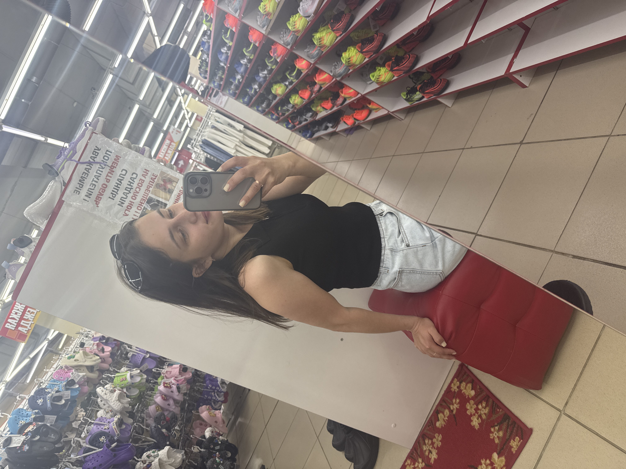 Here is a detailed description of the image:
**Location:** The image appears to be taken inside a shoe store, likely in Chaikovsky, Russia, based on the Cyrillic text visible in the reflection.
**People:** A woman is taking a selfie in the store's mirror. She has dark hair and is wearing a black top and light denim shorts. She is wearing a ring on her finger.
**Objects:** The image includes a mirror, a smartphone used to take the selfie, rows of shoes displayed on shelves, a red cushioned seat, and a small rug with a floral pattern. There is also a black pair of shoes on the floor near the rug.
**Actions:** The woman is taking a selfie, holding her phone in front of her face. She is seated on the red cushion.
**Interactions:** The woman is interacting with the environment by taking a photo of herself within the store.
**Environment:** The environment is a shoe store with well-lit aisles and displays. The floor is tiled. The shoe shelves are stacked with various athletic shoes and sandals, and the red seat and rug are located near the mirror.