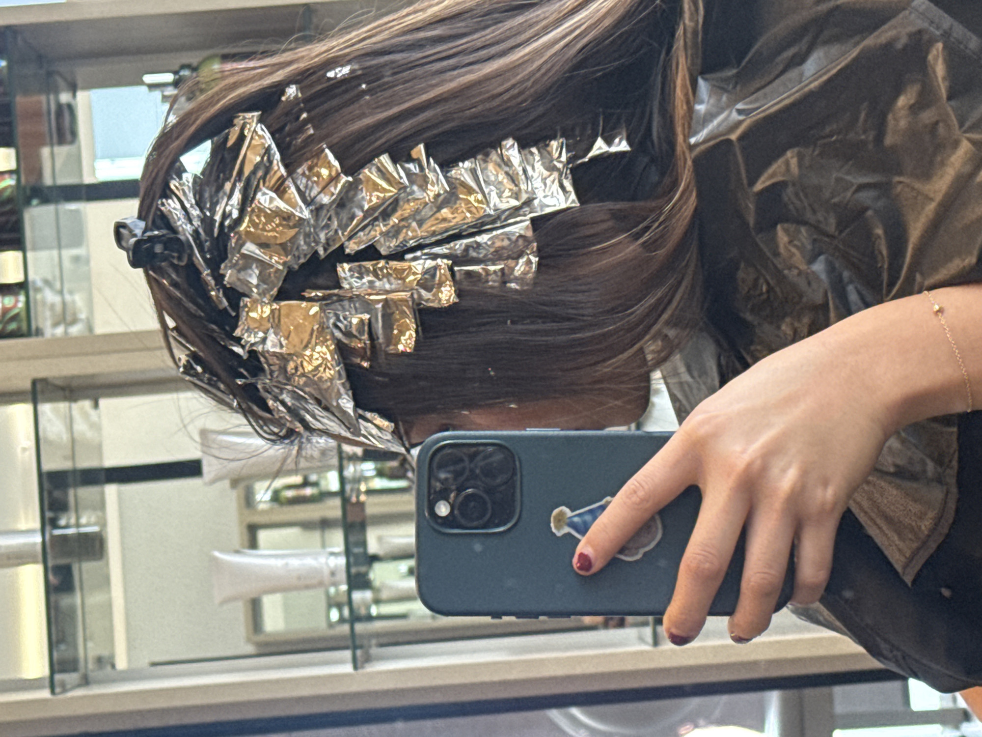 A person with brown hair, partially covered in silver foil highlighting treatments, is taking a selfie in a mirrored salon setting. The individual is wearing a black gown. A blue smartphone with a light-colored sticker is visible in their hand. The background shows shelves stocked with haircare products, reflected in the mirror. The location is a hair salon in an unidentified city.