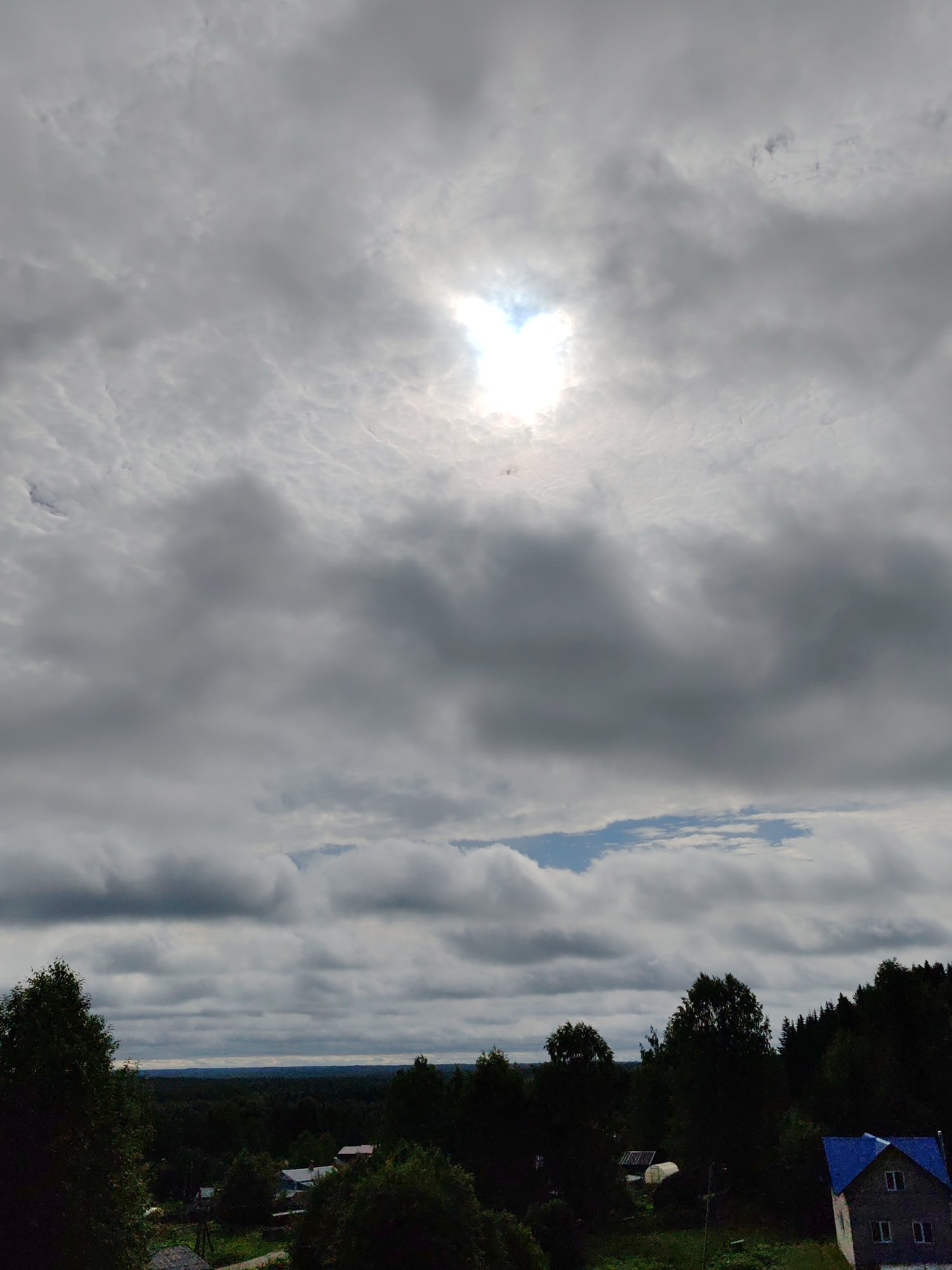 The image depicts a rural landscape in the city of Chukhlem, Russia, dominated by a dramatic sky filled with heavy, gray clouds. A bright spot of sunlight pierces through the cloud cover near the top center of the frame, creating a halo effect. Below the sky, a dense forest stretches across the horizon, with the tops of trees creating a dark, undulating line. In the mid-ground, several small houses with visible roofs are nestled amongst the trees. One house, to the right, stands out with a bright blue roof. The foreground is occupied by the dark silhouettes of trees. The scene conveys a somber, overcast atmosphere typical of a cloudy day in a rural setting. There are no people or animals visible in the image.