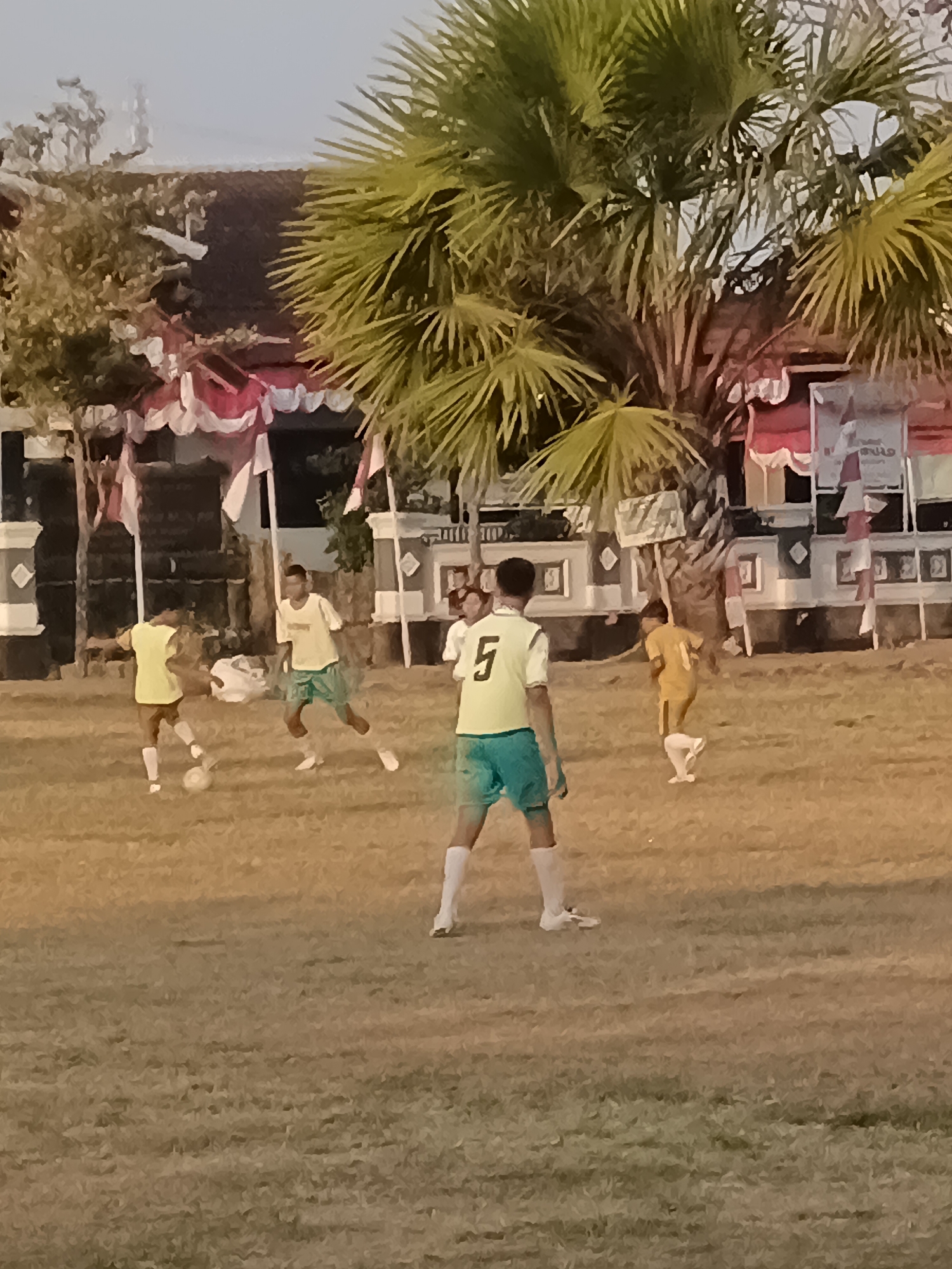 The image depicts a group of young men playing soccer on a grassy field in Desa Gunungsimping, Indonesia. The game appears to be informal, with players wearing simple athletic attire. Two individuals in yellow shirts and green shorts are prominently featured, one of whom is kicking a soccer ball. Another player in a yellow shirt and white shorts is visible further in the background, along with a player wearing a yellow and white jersey with the number "5" on the back. The environment is characterized by dry, yellowish grass, suggesting it may be a warm climate. In the background, there are buildings adorned with red and white banners, possibly indicating a local event or celebration. Tall palm trees with fan-shaped fronds are scattered throughout the scene, adding to the tropical ambiance. The sky is a pale, hazy blue, and the overall lighting suggests it is either early morning or late afternoon. The presence of flags and decorations implies a community gathering or sporting event taking place in the village.