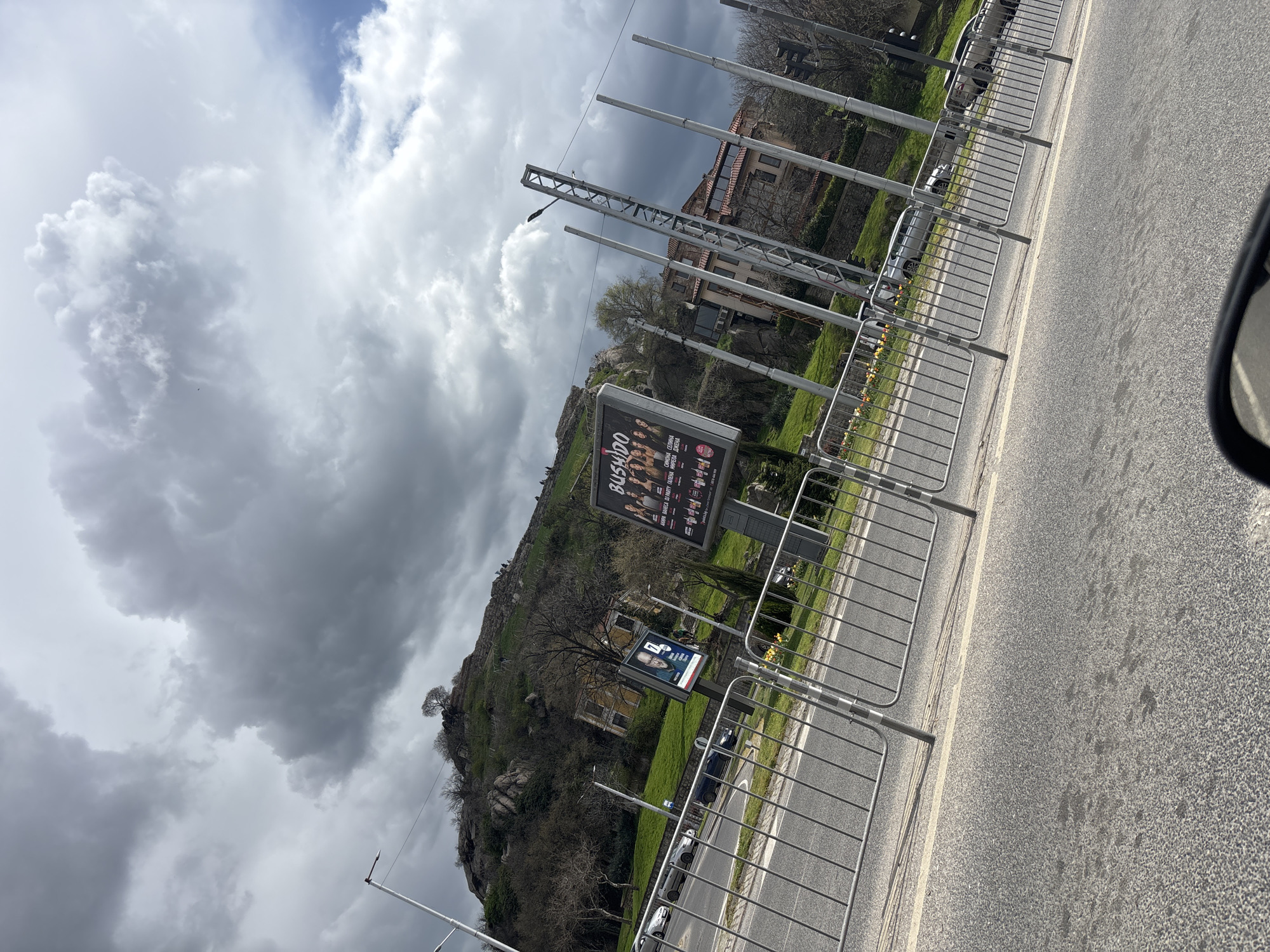 The image shows a street in Plovdiv, Bulgaria. The road is asphalt and has a white dividing line. A metal railing runs along the side of the road. On the other side of the railing, there is a grassy slope leading up to a rocky hill. There are buildings on the hill, including what appears to be a house. There are two billboards near the road. The billboard shows a group of people and the word "BUSHIDO". The sky is cloudy.