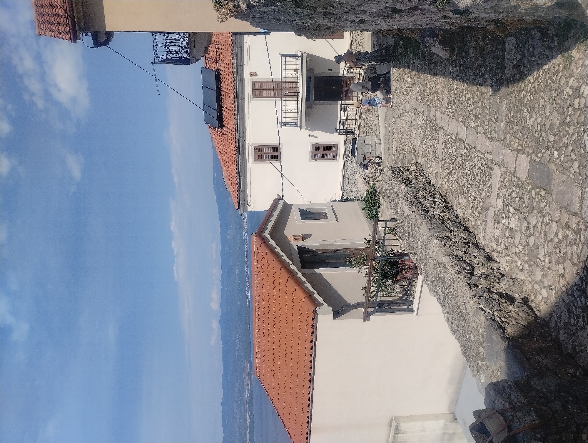 A bright, sunny day shows a cobblestone pathway leading downhill in a village, likely in Općina Vrbnik, Croatia, given the characteristic architecture and coastal scenery. On the left, a cream-colored building with a terracotta tile roof features a balcony with a metal railing and potted plants. Further up the path, a larger white house with a similar terracotta roof has solar panels installed on it. A wrought-iron balcony with decorative scrollwork juts out from this house. Several people are visible on the path. In the middle distance, a man and a young child walk down the path, with another person in the background. To the right of the path, a stone wall forms the boundary of a property, with vegetation growing on it. The backdrop includes a body of water and distant hills, suggesting a coastal setting.
