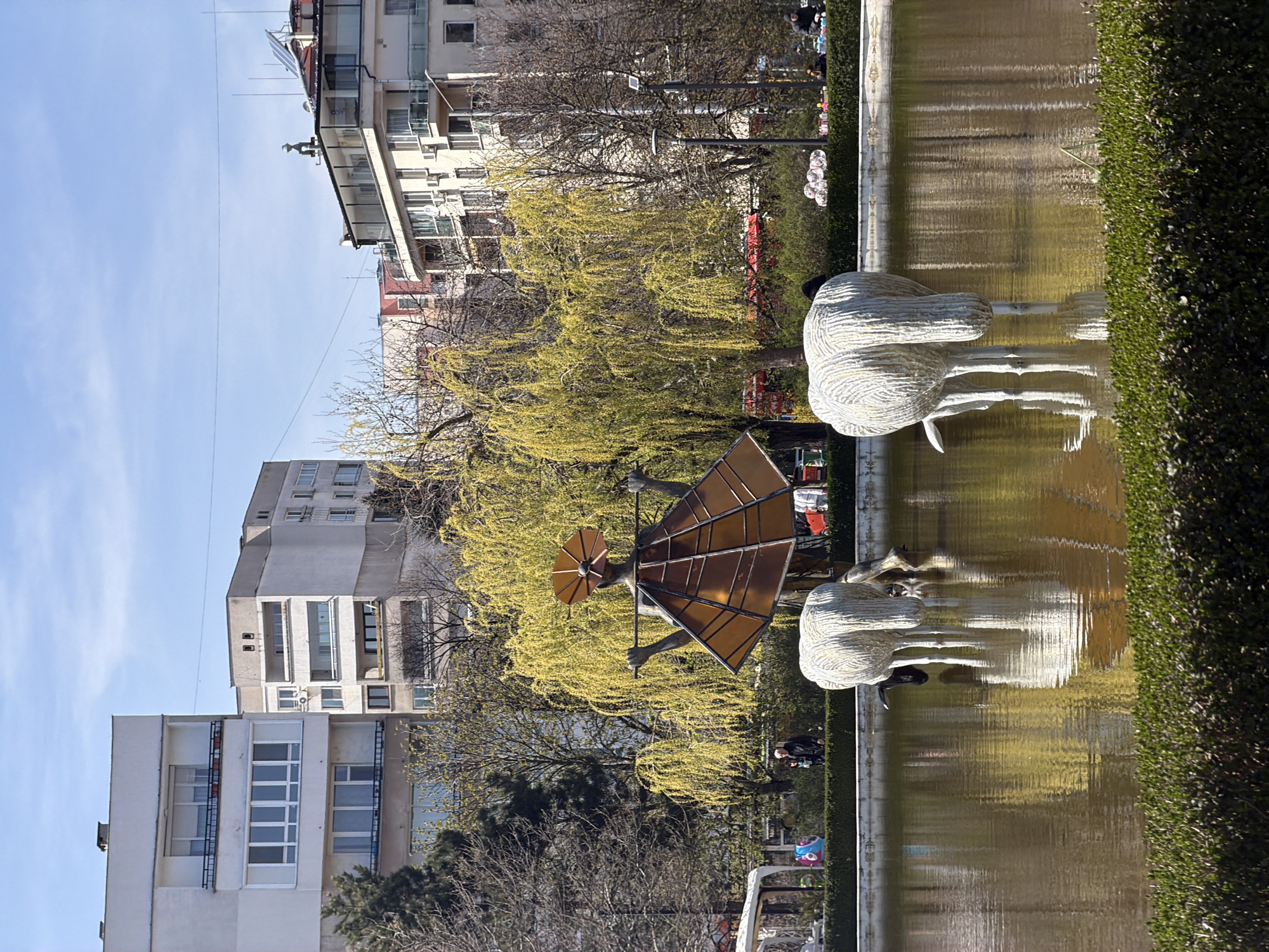 The image depicts an urban park scene in Sofia, Bulgaria, featuring a long rectangular pond with a hedge along one side. In the foreground, two abstract sculptures of birds stand on slender legs at the edge of the pond, their reflections visible in the water. Further back, another sculpture of a man holding a sun-like disc is visible amidst the trees with yellow blossoms.
Behind the pond and sculptures are trees, with residential buildings forming the backdrop of the scene. People can be seen walking in the park. The sky is partially visible and appears clear.