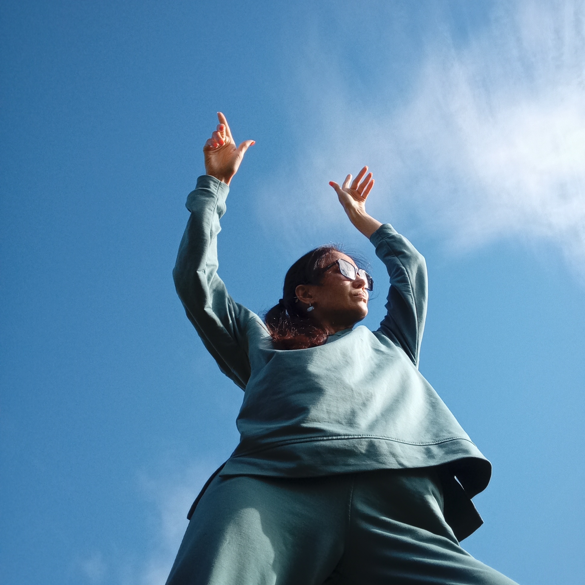 A low-angle shot captures a woman from the chest up, silhouetted against a clear blue sky. She is wearing a teal-colored sweatshirt and pants, with her arms raised high above her head. Her hands are open, palms facing upwards, as if reaching for the sky. She is wearing large, dark-rimmed sunglasses that reflect the bright sky. Her hair is pulled back in a ponytail, and she has a serene expression on her face. A few wisps of white clouds are visible in the upper right corner of the frame, suggesting a bright, possibly sunny day. The perspective makes the woman appear grand and heroic against the vast expanse of the sky. There is no visible text. The setting appears to be outdoors, possibly in a natural environment, given the ample sky and the woman's relaxed attire. The location context provided is Burenka, Russia.