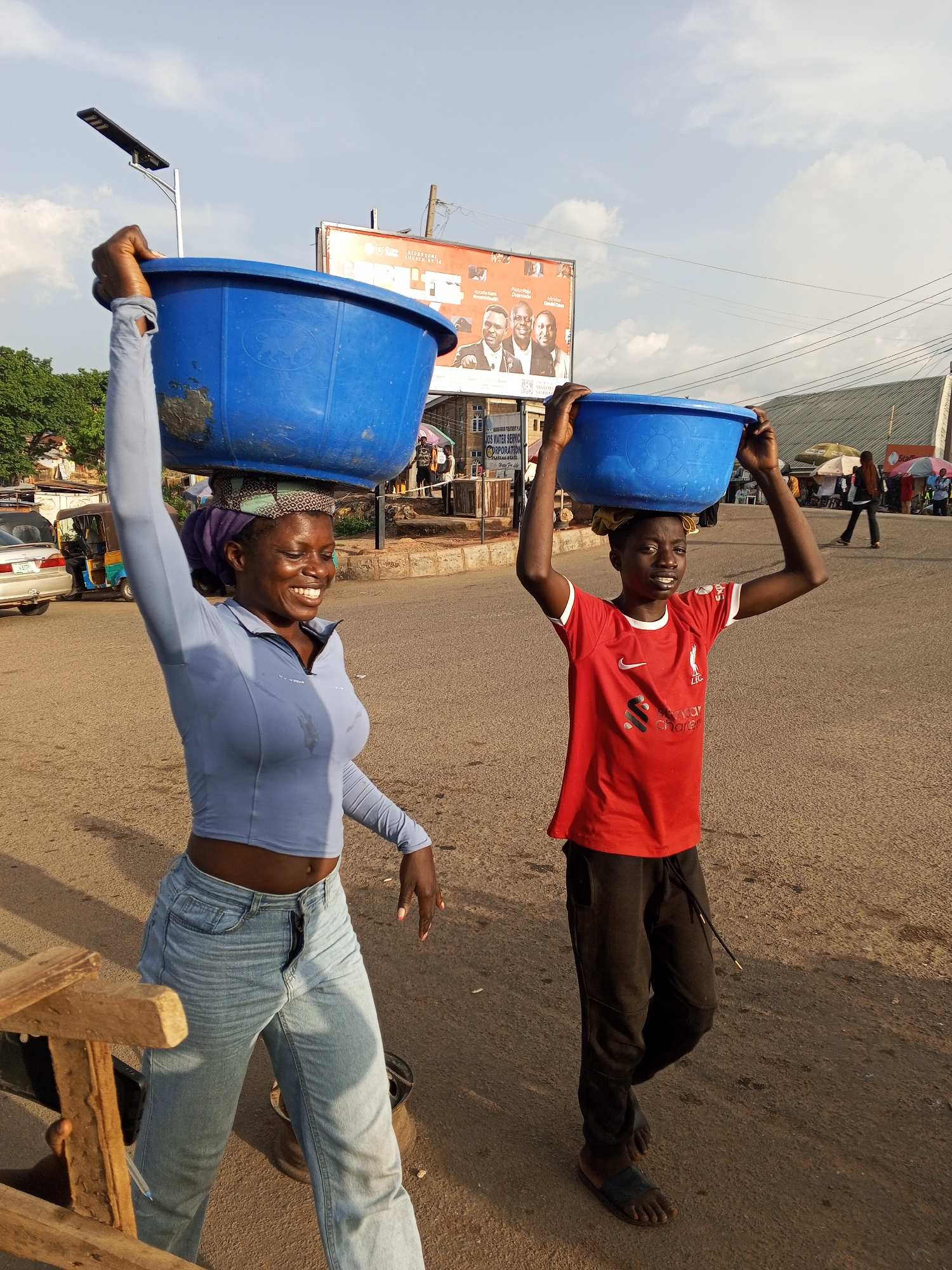 Two people are carrying blue plastic tubs on their heads on a street in Gwong, Nigeria. The person on the left is a woman with a purple headscarf, wearing a light blue cropped long-sleeve shirt and light-wash jeans. Her right arm is raised. The person on the right is a young boy wearing a red Liverpool soccer jersey and black sweatpants with sandals. Both individuals are smiling. In the background, there is a billboard, a street lamp, trees, and other people. A car and a "Keke Napep" (auto rickshaw) are parked on the left side of the street. There is also some sort of wooden structure in the lower left of the frame. The sun appears to be setting.