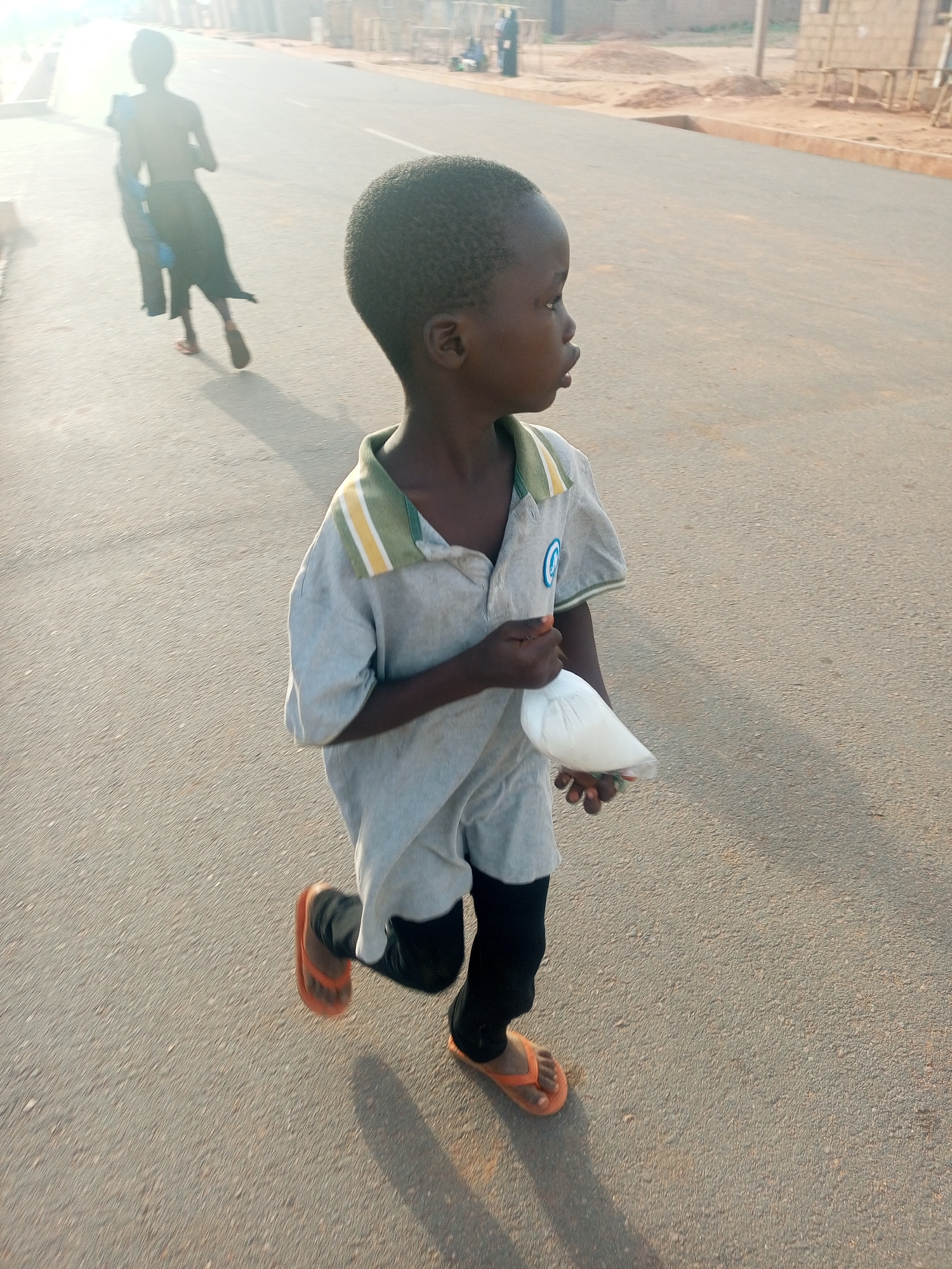 A young boy walks along a paved road in Tempure, Nigeria. He wears a short-sleeved, light gray shirt with a green and yellow striped collar, black leggings, and orange flip-flops. He holds a white object, possibly food in a bag, in his hands. Two figures, appearing to be children, are visible in the background, walking away from the viewer on the same road. The road appears to be in a residential area with buildings visible in the background. The lighting suggests it is daytime.