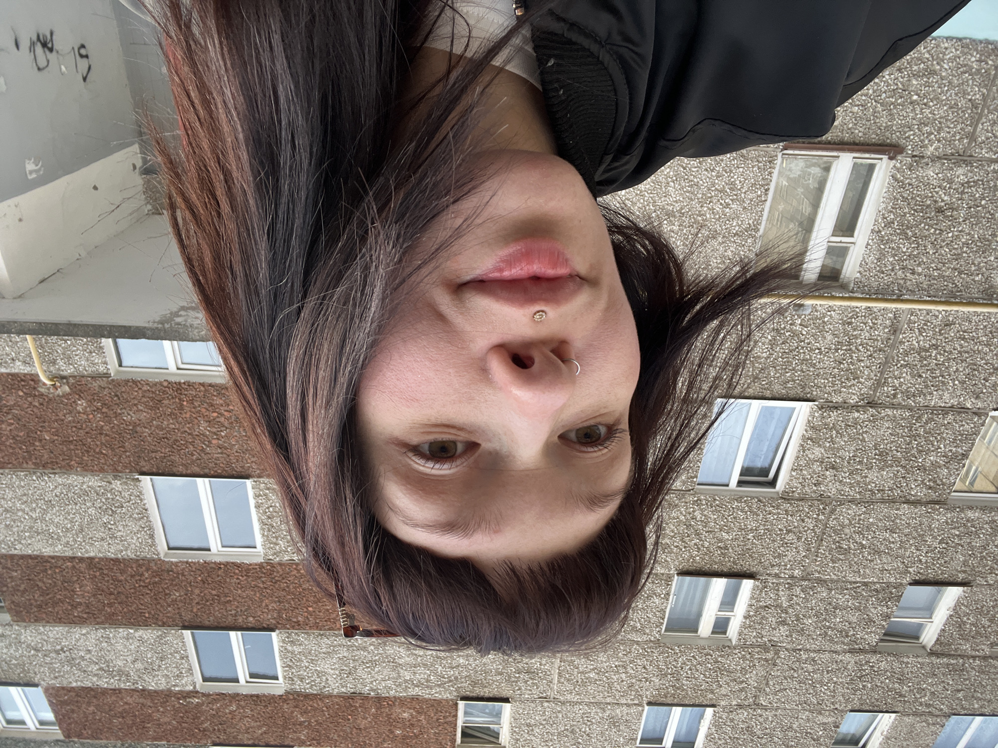 A female subject is depicted in a slightly low-angle, close-up photograph taken in an urban environment identified as Izhevsk, Russia. She has dark brown hair with reddish-brown highlights, cut with bangs, and a subtle smile. She presents a silver hoop piercing in her septum and a gold-colored labret stud on her philtrum. Her attire includes a black bomber-style jacket over a white garment.
The background consists of a multi-story residential building, likely a panel construction from the Soviet era. Its facade is characterized by a coarse, textured aggregate finish (pebble dash) in alternating horizontal bands of light grey and reddish-brown. Numerous white-framed windows are visible across the building's exterior. A yellow utility pipe is horizontally affixed to the building in the lower-mid right. The bottom right of the image shows a partial view of a concrete architectural element, possibly a balcony or entrance overhang, with black graffiti. The lighting indicates an overcast sky.