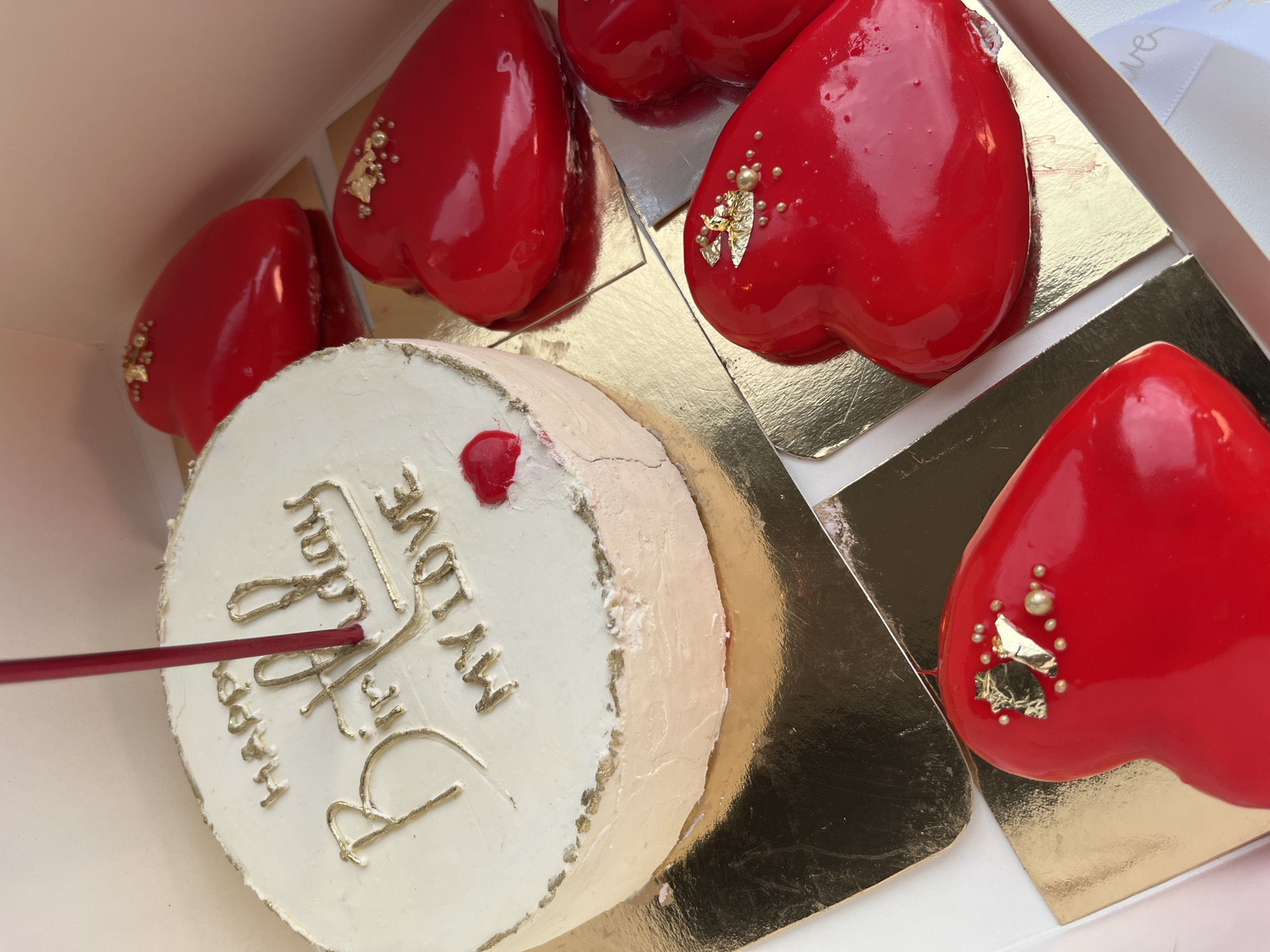 The image shows a collection of glossy, red, heart-shaped pastries and a round cake in a box. The pastries are decorated with gold leaf and small, pearl-like embellishments. The cake is white with gold lettering spelling out "Happy Birthday My Love". A red stick is inserted into the cake. The pastries and cake are sitting on gold-colored squares in a white box. The location appears to be Unknown, Unknown.