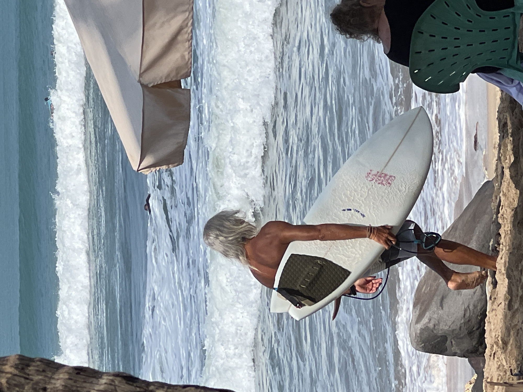 A surfer with long, grey hair carries a white surfboard towards the ocean in Kuta, Indonesia. The surfer's back is to the camera, and they are wearing dark swim trunks. The surfboard has a dark grip pad on the tail and a red decal. In the background, waves crash onto the shore, and other surfers are visible in the water. A beige beach umbrella is partially visible in the upper right corner, and two people are seated in green chairs on the sand to the right. Large rocks are present on the beach. The scene depicts a sunny day at the beach, with clear blue skies and moderate waves.