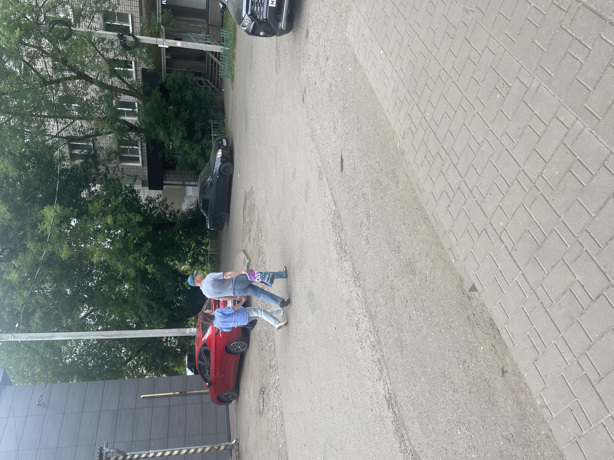In Zhukovo, Russia, on a cloudy day, two individuals, a woman and a man, are walking across an asphalt road in front of a building with a dark grey facade and a large tree. The woman, wearing a blue shirt and light-colored jeans, walks slightly ahead of the man. The man, in a grey polo shirt, blue jeans, and a blue baseball cap, carries a colorful shopping bag. A red car is parked to their left. To the right, a black sedan is parked, and a portion of a black SUV is visible in the foreground. The ground is a mix of asphalt and paving stones.