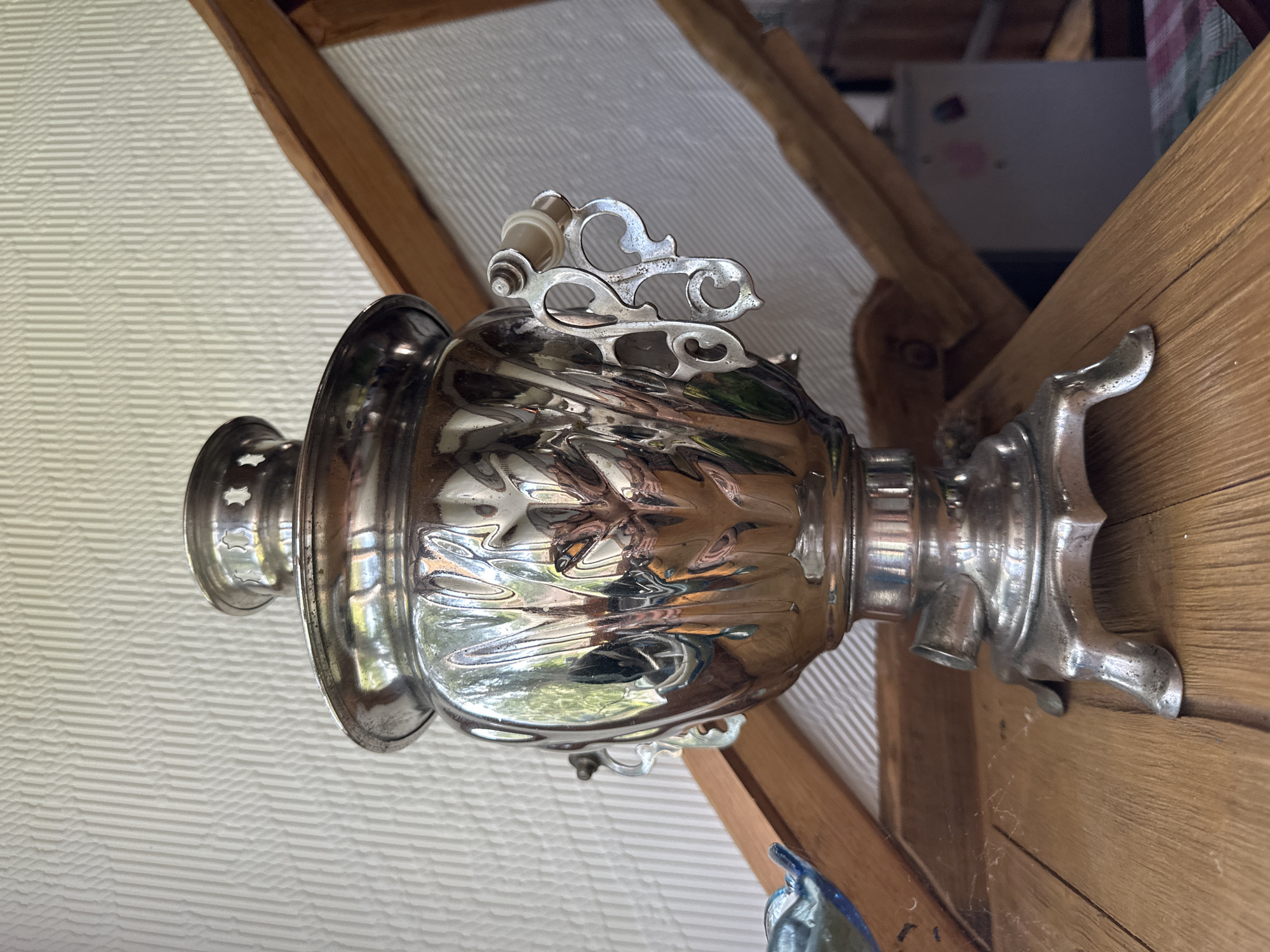 A close-up, eye-level shot focuses on a decorative, silver samovar resting on a wooden surface, possibly a table. The samovar is ornate, with intricate carvings and handles, and its polished surface reflects the surrounding environment, including greenery and wooden structures. The background features a textured, off-white wall and a wooden beam structure, suggesting an indoor or covered outdoor setting. The scene is lit by natural light, creating highlights and shadows on the metallic surface of the samovar. The image appears to be taken in Norviliškės, Lithuania, specifically within the grounds of the Norviliškės Castle.