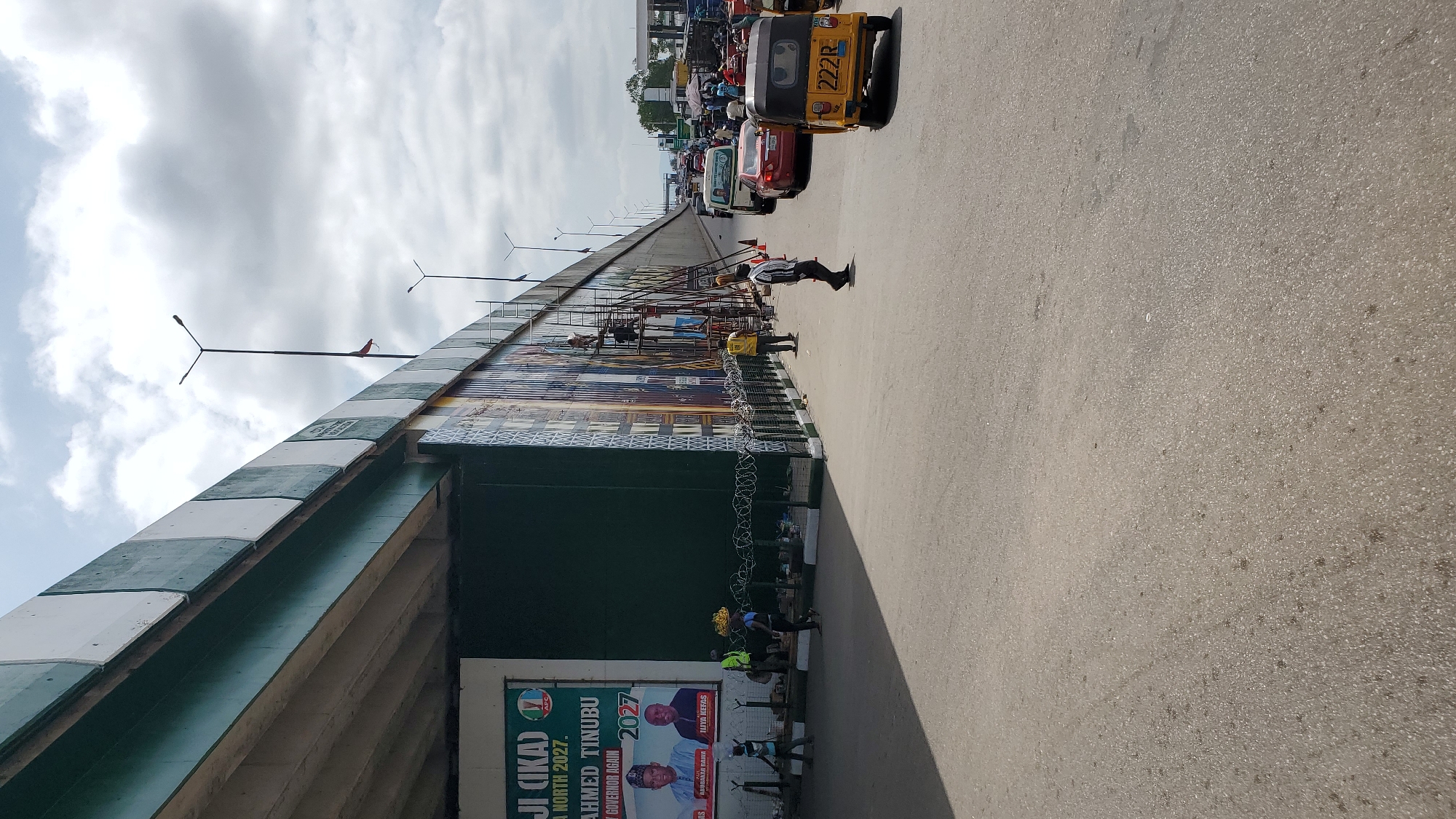 This is a street-level, landscape-oriented photograph taken from the side of a road in Jalingo, Nigeria. The image is rotated 90 degrees counter-clockwise, so the top of the scene is to the left. A busy road is visible with traffic, including a yellow motorized tricycle with "222R" on its back, cars, and other vehicles.
Along the left side of the road is a pedestrian overpass or bridge, adorned with a large mural and decorative tiles. The overpass has lights mounted on poles extending from its side. Several people are working on or near the overpass, with scaffolding visible. Some are dressed in construction attire, while others appear to be passersby. Barbed wire fencing is present along the base of the overpass.
At the bottom of the overpass, a large political banner is displayed. The banner features images of two men and text, including "AHMED TINUBU" and "GOVERNOR AGAIN 2027." The presence of the APC logo suggests this is a campaign banner for the All Progressives Congress.
The sky above is partly cloudy with white clouds against a pale blue background, indicating daytime. The road surface is asphalt. The overall scene depicts a vibrant urban environment with ongoing construction and political activity.