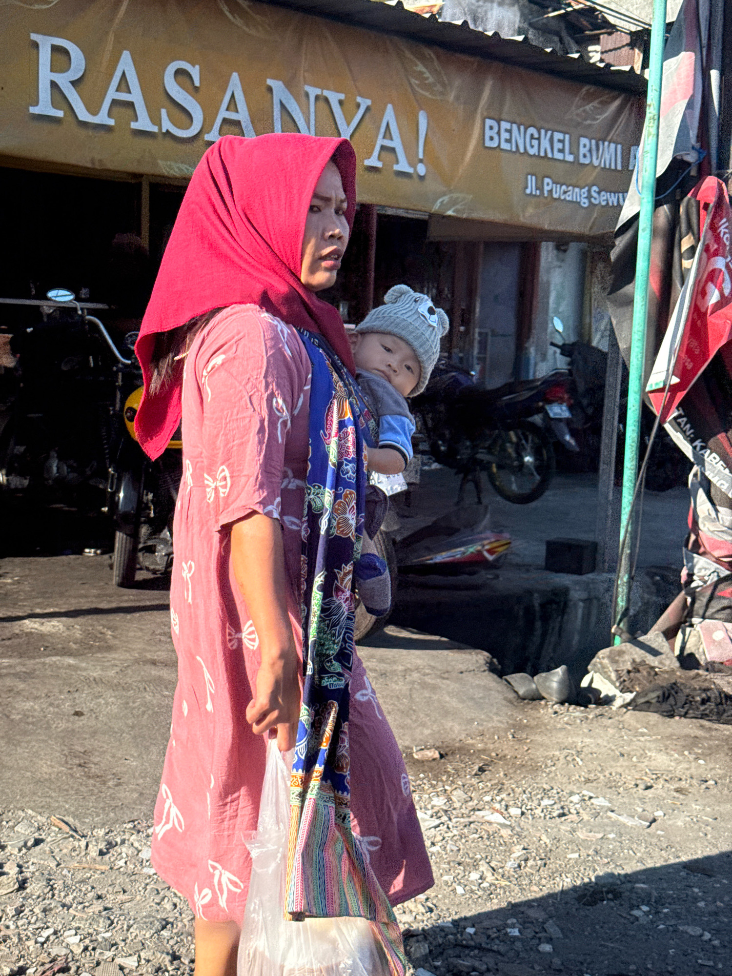 A woman wearing a pink dress and a red headscarf carries a baby in a sling. The baby is wearing a grey hat with bear ears. The woman is walking on a dirt road next to a building with a sign that reads "RASANYA!" and "BENGKEL BUMI Jl. Pucang Sewu". There are motorcycles parked in the background and a green pole with flags attached to it on the right side of the image. The scene appears to be a street in Surabaya, Indonesia.
