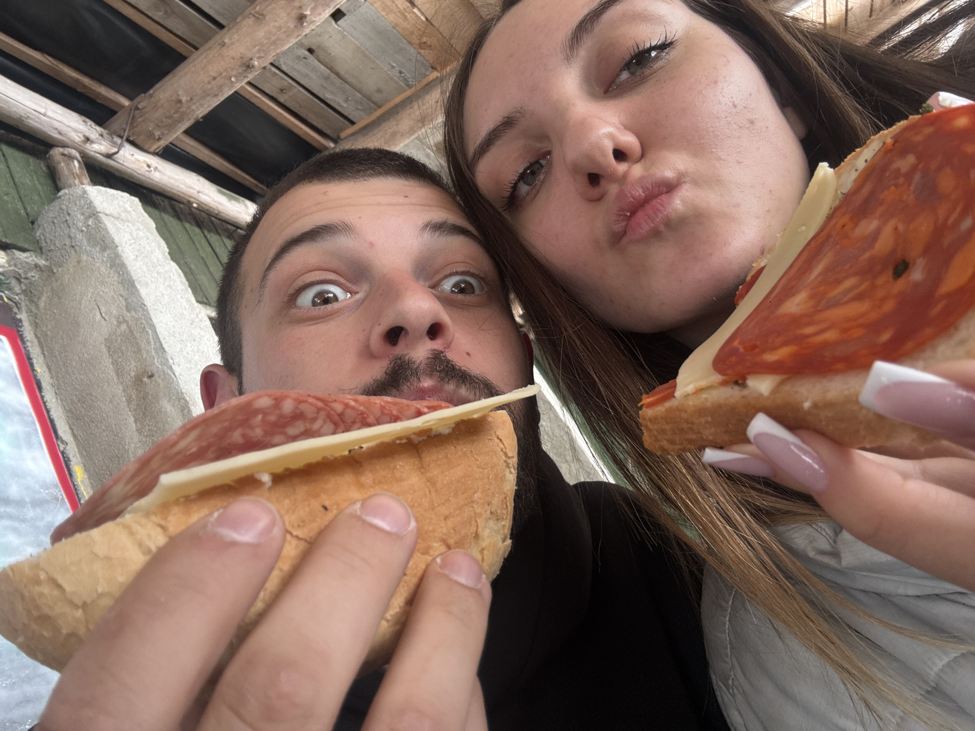 This close-up, low-angle selfie features a man and a woman humorously posing with their sandwiches. On the left, a man with short dark hair, a mustache, and light stubble gazes wide-eyed at the camera, conveying a comical, surprised expression. He holds a substantial piece of baguette-style bread topped with multiple slices of what appears to be salami or cured sausage and a single slice of light-colored cheese.
On the right, and slightly closer to the viewer, a woman with long, straight, light brown hair pouts her lips into a "kissing face." Her eyes are also directed towards the camera. She holds a similar sandwich, featuring the same bread, salami, and cheese. Her hands reveal long nails, elegantly styled with a white French tip manicure.
The background provides context for the casual setting, showing a ceiling structure composed of rough wooden beams, some dark paneling, and a weathered concrete or stone pillar on the left. This rustic environment suggests an outdoor or semi-covered eating spot, possibly a local eatery or market stall in Sofia, Bulgaria, where the pair is enjoying a casual snack. The overall atmosphere is playful and candid. There is no discernible text in the image.