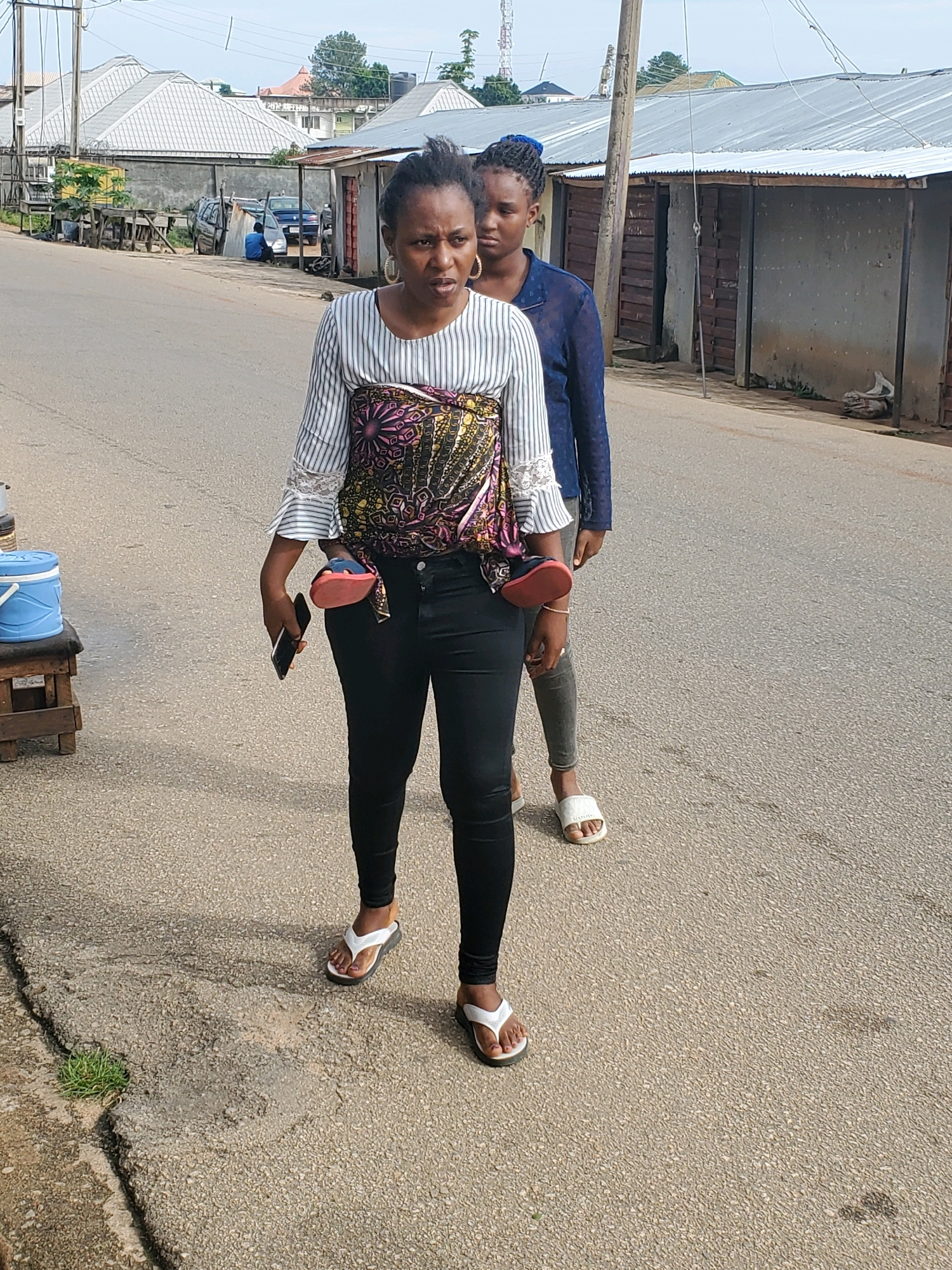 Two women are walking on a paved road in what appears to be a commercial area. The woman in the foreground is wearing a striped long-sleeved shirt with bell sleeves and black pants. She is holding a black smartphone in her right hand and a pair of red flip-flops in her left hand. The second woman, slightly behind and to her right, is wearing a dark blue long-sleeved shirt and grey pants, with white flip-flops. The environment includes buildings with corrugated metal roofs, a wooden structure with a blue bucket on it, and a grey concrete wall. The road shows signs of wear and tear, with cracks and some vegetation growing in them. The background shows more buildings and what might be utility poles and wires, suggesting an urban or semi-urban setting.