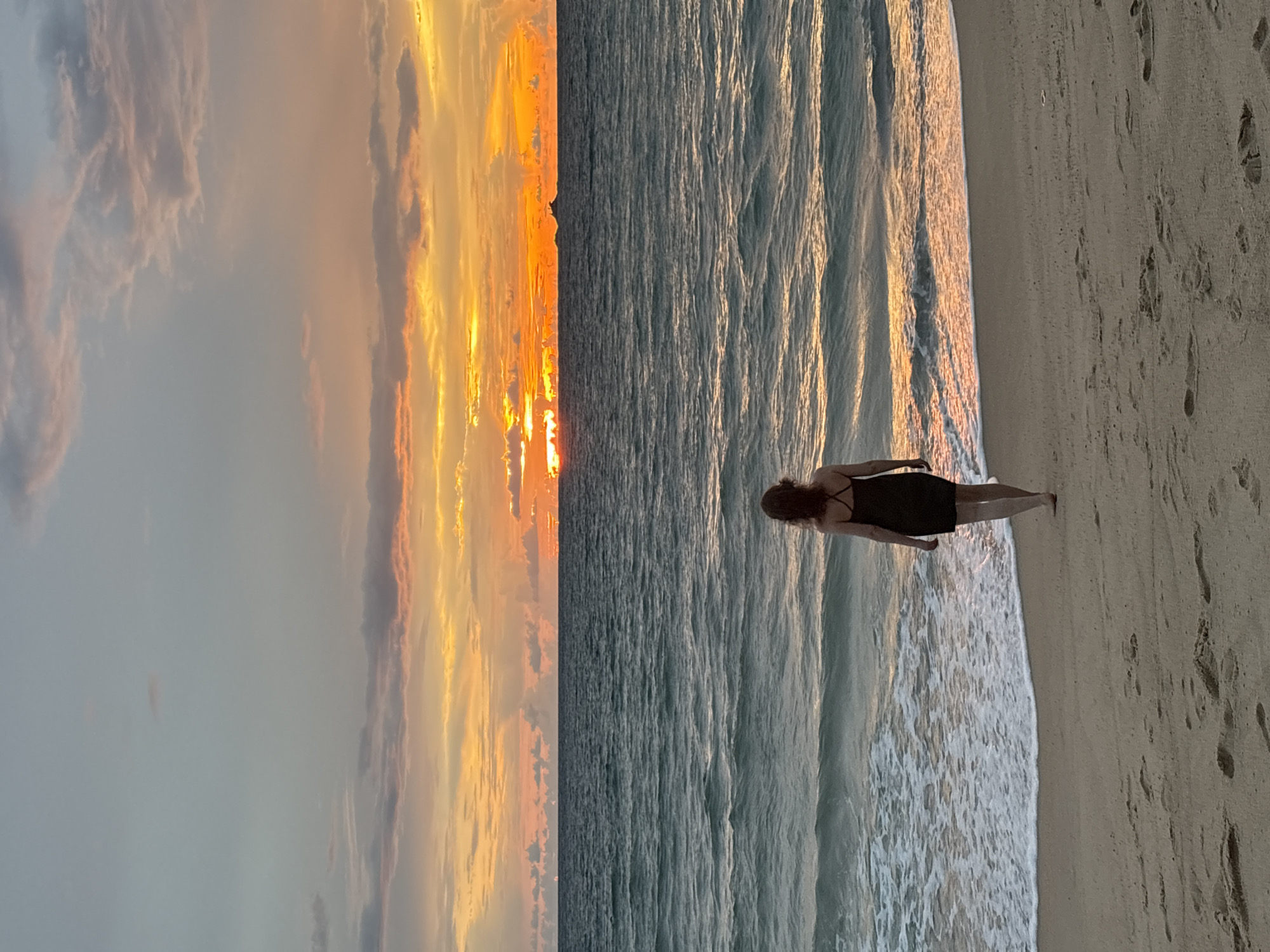 A woman stands on a sandy beach, facing the ocean. The sun is setting, casting a warm glow on the sky and the water. The waves are gently rolling onto the shore. The woman is wearing a dark-colored dress and her hair is pulled back. Her posture suggests she is looking out at the sunset. The sand is marked with footprints. The scene evokes a sense of peace and tranquility. The time of day is sunset. The weather appears to be calm and pleasant. The location is a beach, likely in Choeng Thale, Thailand. There is no visible text in the image.