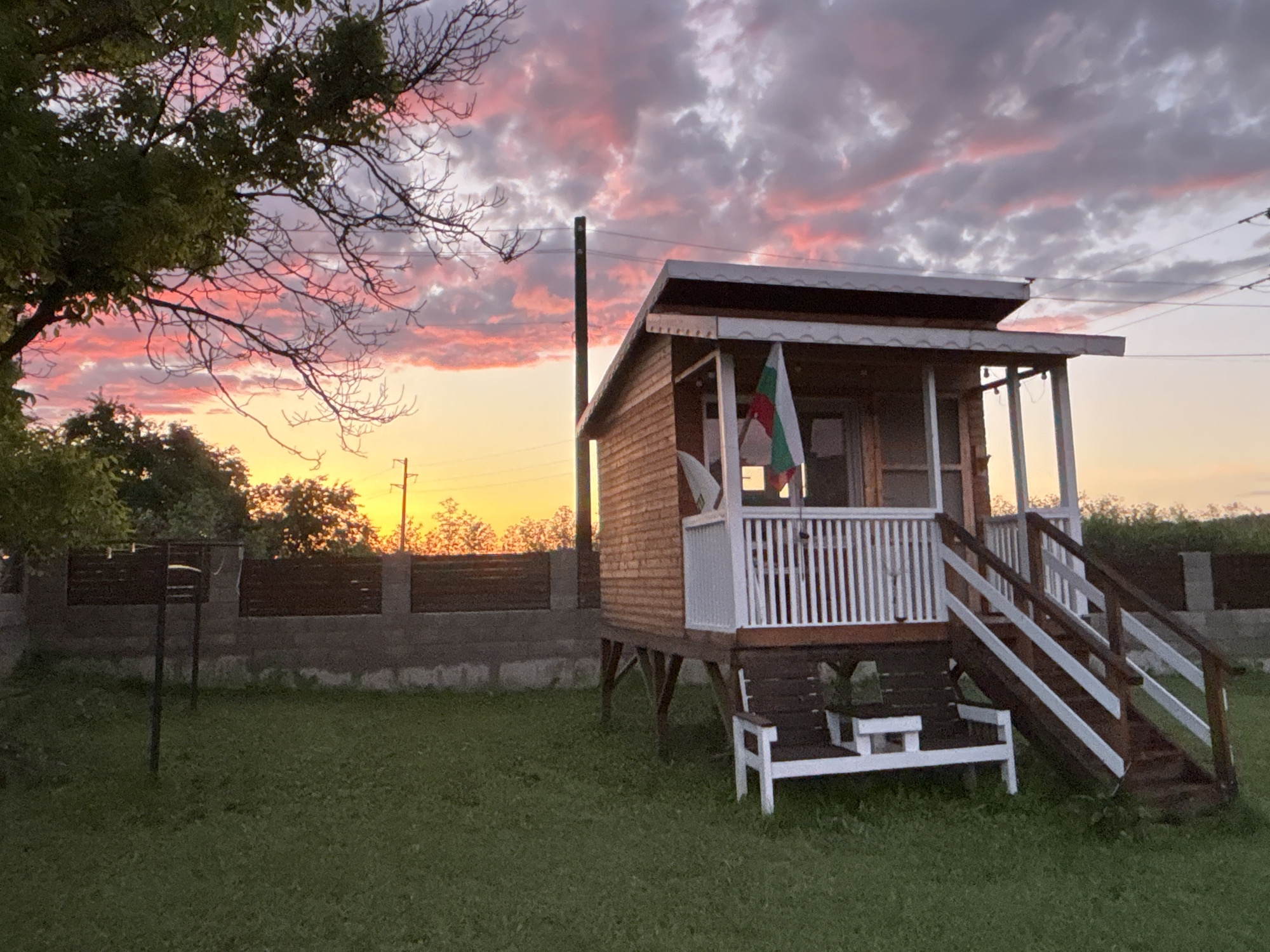 This image depicts a serene outdoor scene at sunset in Pchelnik, Bulgaria. The central subject is a small, elevated wooden house with a porch featuring a white wooden railing and a set of matching stairs. A small white wooden bench is positioned below the stairs. A Bulgarian flag, with horizontal white, green, and red stripes, hangs from the porch, providing a clear location cue. A satellite dish is attached to the side of the house.
The foreground consists of a lush green lawn. To the left, a metal frame or stand is visible on the grass. A fence, made of a concrete base topped with dark wooden slats, runs horizontally behind the house and the lawn. Beyond the fence and house, the sky is illuminated by the setting sun, creating a striking palette of vibrant orange and yellow near the horizon, gradually transitioning into soft pink and purple clouds across the upper sky. Silhouetted trees and distant foliage line the horizon. A tall utility pole stands behind the house, with power lines visible stretching across the top right.
The time of day is clearly sunset, evident from the dramatic colors in the sky and the warm, soft light. There are no people visible, and no discernible text. The scene conveys a peaceful and tranquil atmosphere.