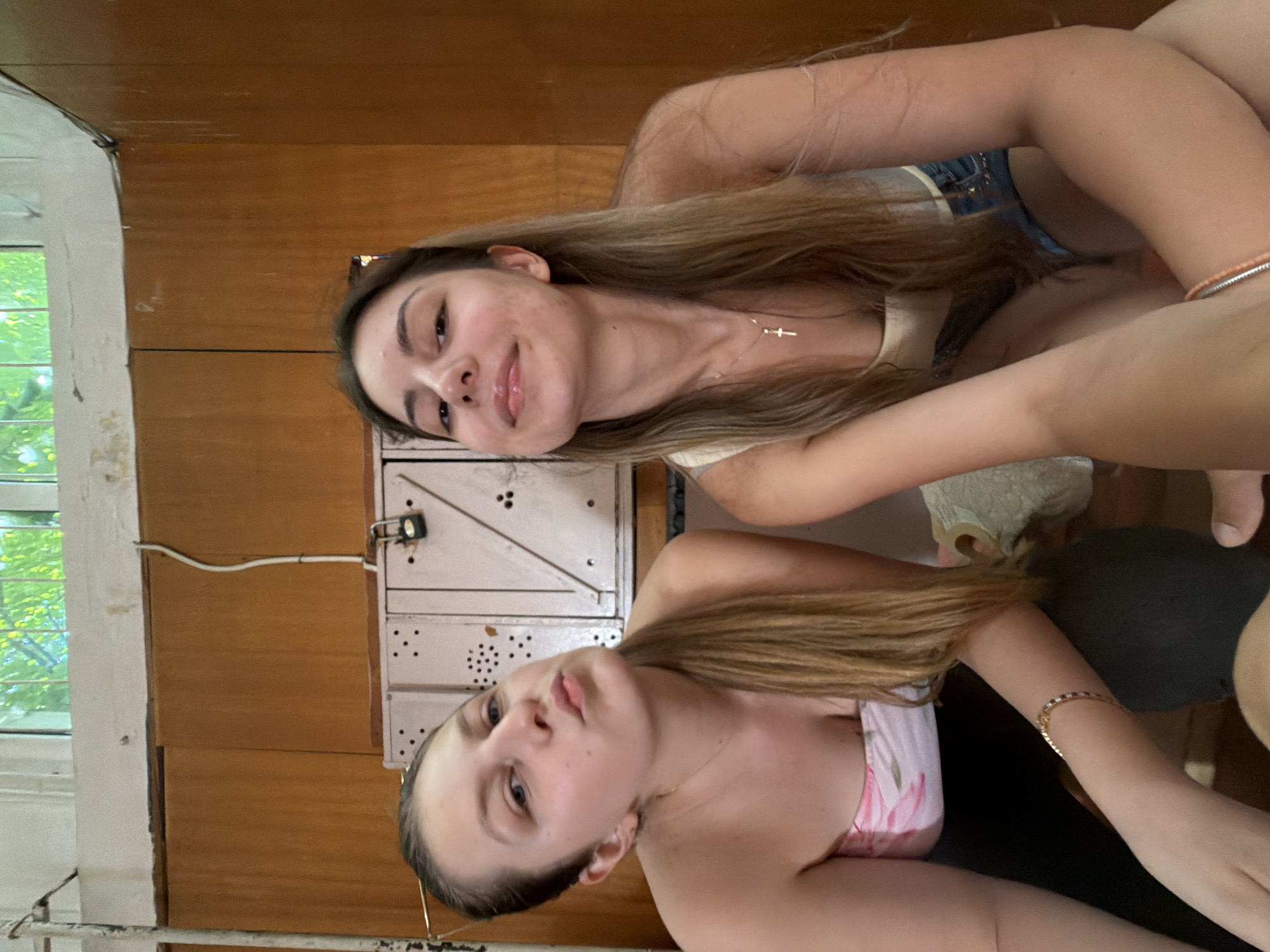 Two young women are posing for a selfie in what appears to be a room with wooden paneling and a locker system in the background. The woman on the left is wearing a strapless pink and white floral top and has her hair pulled back in a ponytail. She is pouting her lips and looking towards the camera. The woman on the right is wearing a light-colored tank top and denim shorts. She is smiling slightly and looking at the camera. Both women have their arms visible, with one woman wearing a bracelet. The locker system is pale pink with black accents and a small padlock on one of the doors. The room has a window at the top with greenery visible outside, suggesting it might be daytime. There is also a white wire running along the wall. The overall setting appears to be indoors, possibly in a locker room or communal area. The image does not provide any specific clues to pinpoint its location within Burgas, Bulgaria.