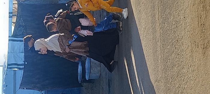 Six individuals—three adults and three children—walk from left to right across an unpaved, dusty area in Khan Yunis, Palestinian Territory. Strong sunlight from the left casts long shadows across the ground.
The group is led by a young girl in a yellow hooded tracksuit and white sneakers. Behind her, a young child in blue jeans walks beside an adult, mostly obscured, who carries a toddler in a brown hooded top on their hip. Next to this adult, a woman in a light-colored hijab, a patterned brown and black outer garment, a dark dress, and a brown tasseled scarf walks with a faint smile, holding a blue object in her left hand. Another woman in a dark hijab is partially visible behind her.
In the background, a blue or white tarp tent is visible on the left. A dark fabric structure, possibly a partition or shelter wall, spans the mid-ground. Above this dark fabric, the head of a young man is visible, looking to the right. Two large, round, metallic containers sit on the ground behind the walking group.
