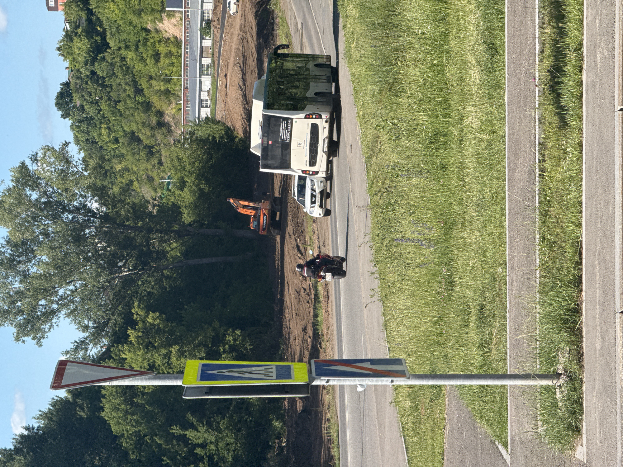 The image shows a street scene with a road, a sidewalk, and a grassy area with trees in the background. On the road, there is a white bus, a white car, and a motorcycle. The bus has advertisements on its rear. The car is driving in the same direction as the bus. The motorcycle is also on the road, with a rider wearing a helmet. There are traffic signs on the side of the road, including a pedestrian crossing sign and a yield sign. In the background, there is a construction site with an excavator and a large building. The sky is blue with some clouds.