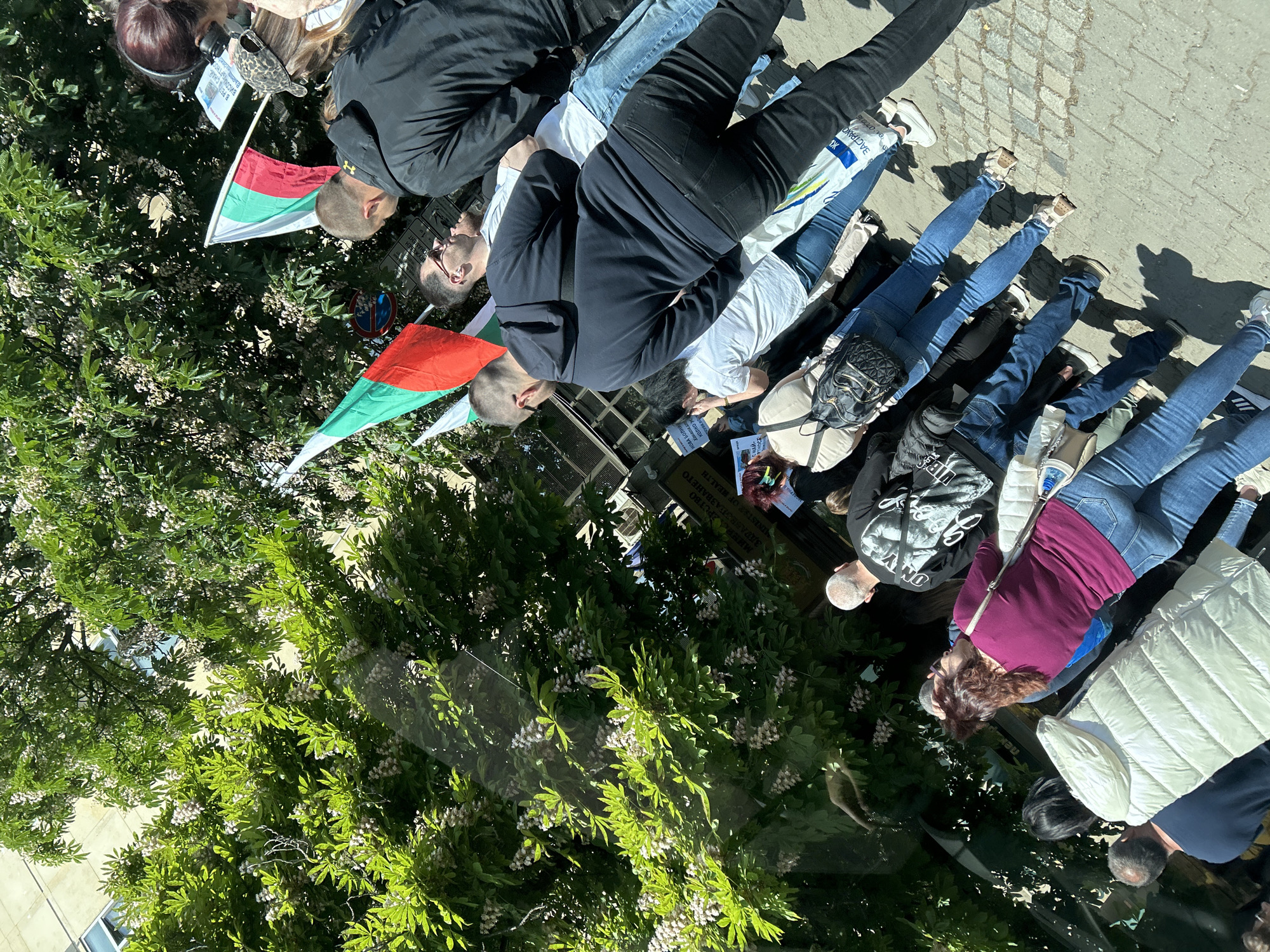 In Sofia, Bulgaria, people are lying on the sidewalk, likely as a protest. Some individuals hold Bulgarian flags, suggesting a nationalistic theme. One person is wearing a maroon shirt, while another wears a white puffy jacket. A sign with what appears to be medical information is visible. The scene is partially obscured by lush green trees, indicating an outdoor setting within the city.