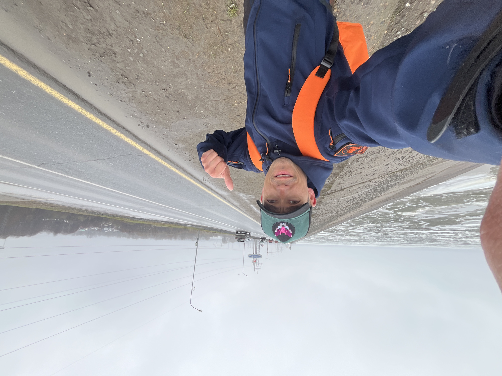 An upright, eye-level selfie shows a man smiling and giving a thumbs-up, standing on the shoulder of a multi-lane road in Noviy, Russia, under an overcast sky. The man is wearing a dark blue and orange jacket with prominent orange shoulder straps and accents, and a dark green baseball cap featuring a pink graphic of a pig's head wearing a gas mask or respirator. He has short dark hair and a clean-shaven face.
To his left is the grey asphalt road with a faded yellow line marking the shoulder. Distant cars are visible on the road, and further down, a large sign structure with illegible Cyrillic text. Several utility poles and power lines crisscross the cloudy sky. To the man's right, a wide, still body of water stretches into the distance, its surface reflecting the grey sky, suggesting a large lake, river, or extensive flooding. The opposite bank of the water features a thin strip of distant trees and some small, light-colored structures. The overall weather appears to be overcast and dull, suggesting a cool daytime. The man's cheerful expression contrasts with the muted, grey tones of the environment.