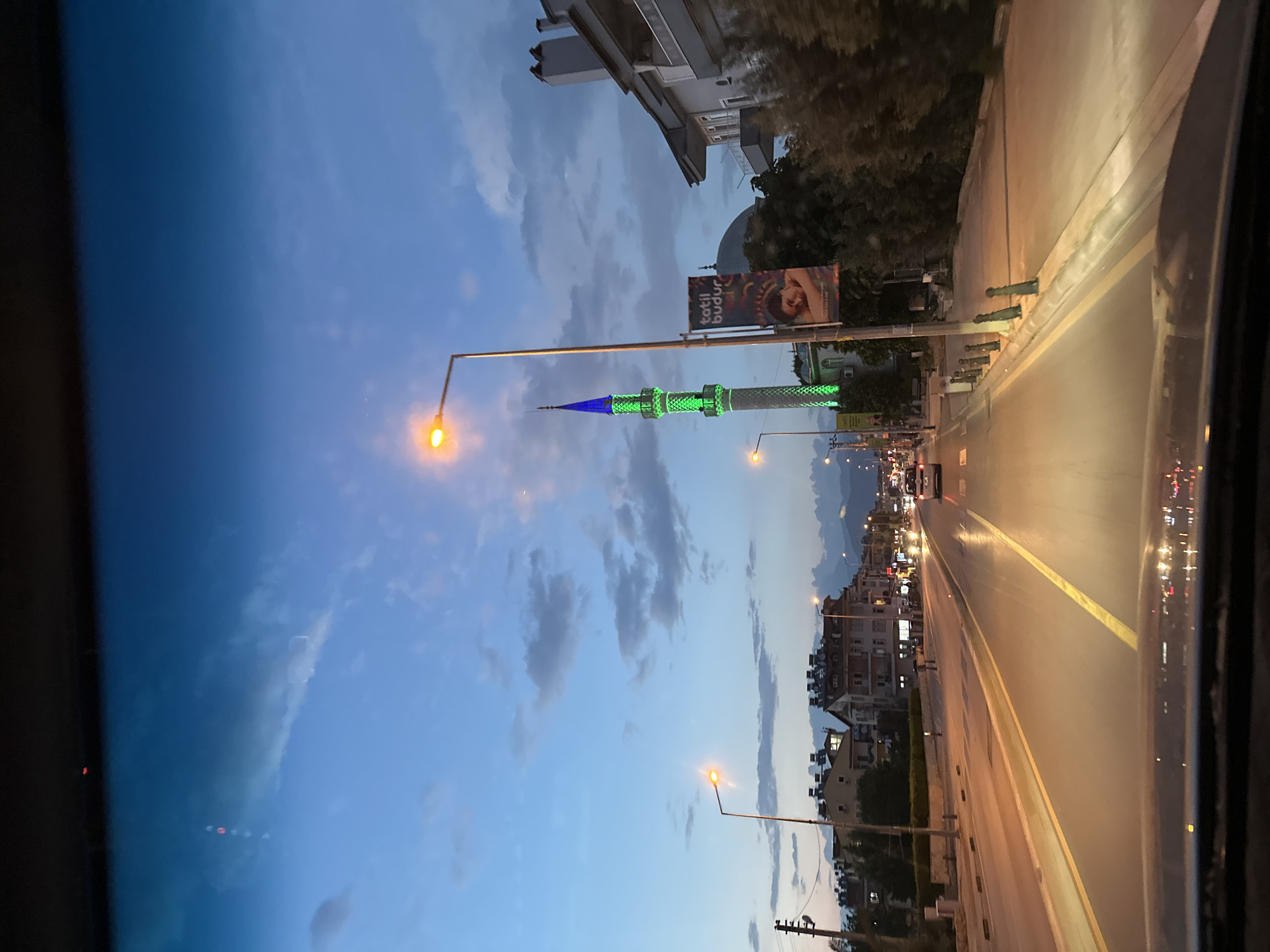 View from a vehicle on a road in Fethiye, Turkey, at dusk. The scene includes a brightly lit mosque minaret, painted with green and blue lights, standing prominently along the roadside. Streetlights illuminate the road, while buildings and mountains are visible in the background. A billboard is placed near the mosque. Cars are driving on the road. The sky is a blend of blue and gray clouds.