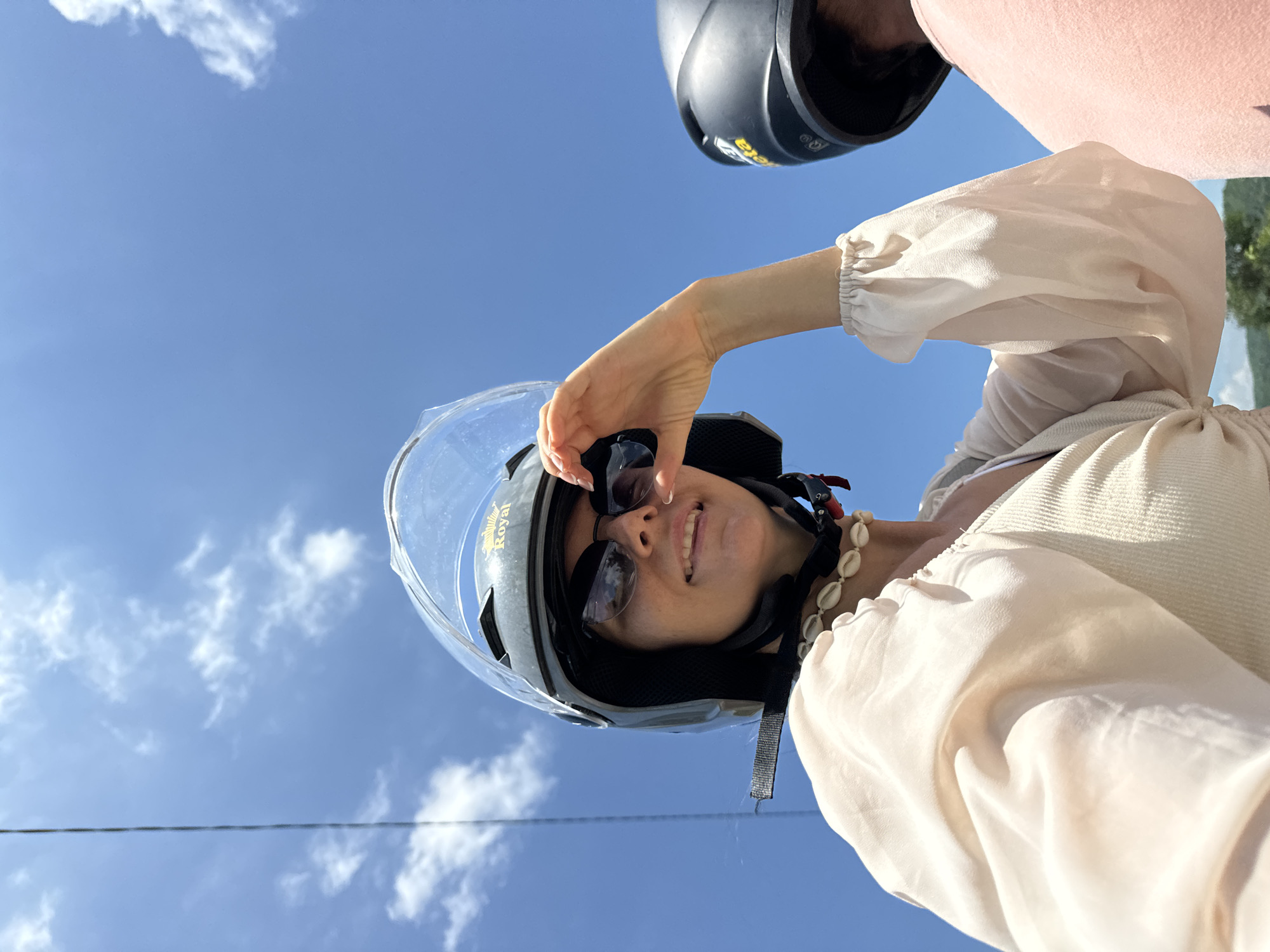 In a low-angle, close-up shot taken in Ham Thuan Bac, Vietnam, a woman wearing a black and grey motorcycle helmet with a clear visor smiles and shields her eyes with her right hand. She has dark sunglasses on and a necklace made of small white shells. She is wearing a light-colored, possibly cream-colored, blouse with puffed sleeves over a ribbed tank top. The sky is bright blue with scattered white clouds. A black helmet and a pink garment are visible in the background to the right, suggesting another person is present. The image appears to be a selfie taken while riding a motorcycle.