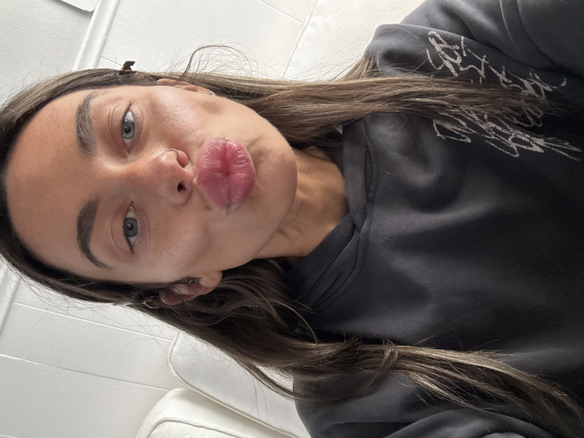 A young woman is captured in a close-up selfie, with her head tilted to the right and her body angled. She makes a playful puckered "kiss" face towards the camera. Her long, dark brown hair cascades over her shoulders and part of her dark grey or black hooded sweatshirt. She has striking blue-grey eyes, natural eyebrows, and a small silver hoop nose ring on her left nostril. A small, dark hair clip is visible in her hair above her right ear. The sleeve of her sweatshirt features abstract white graphic designs, resembling stylized scribbles or unreadable text. The background suggests an indoor setting, with a plain, light-colored wall and what appears to be white or off-white upholstered furniture, possibly a couch or bed, partially visible. The overall mood conveyed is casual and lighthearted. No specific time of day or external location cues are discernible from the image itself.
