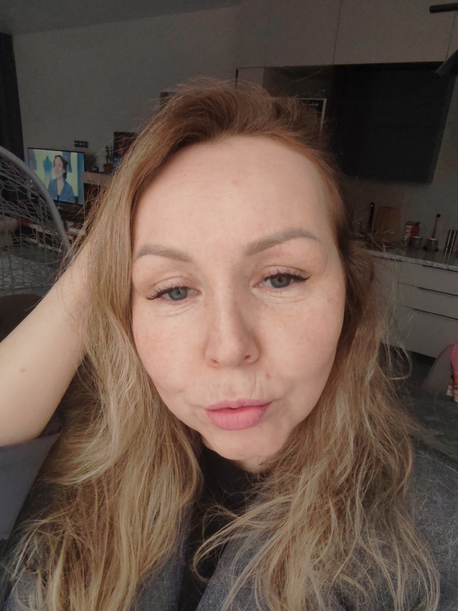A woman with fair skin and light brown hair poses for a selfie indoors. She has light eyes and is making a kissing face. Her left hand is raised and appears to be touching her head. In the blurred background, there is a television displaying a woman, a hanging chair, kitchen cabinets, and various items on the counter. There is no information to suggest the image was taken in Chaikovsky, Russia.