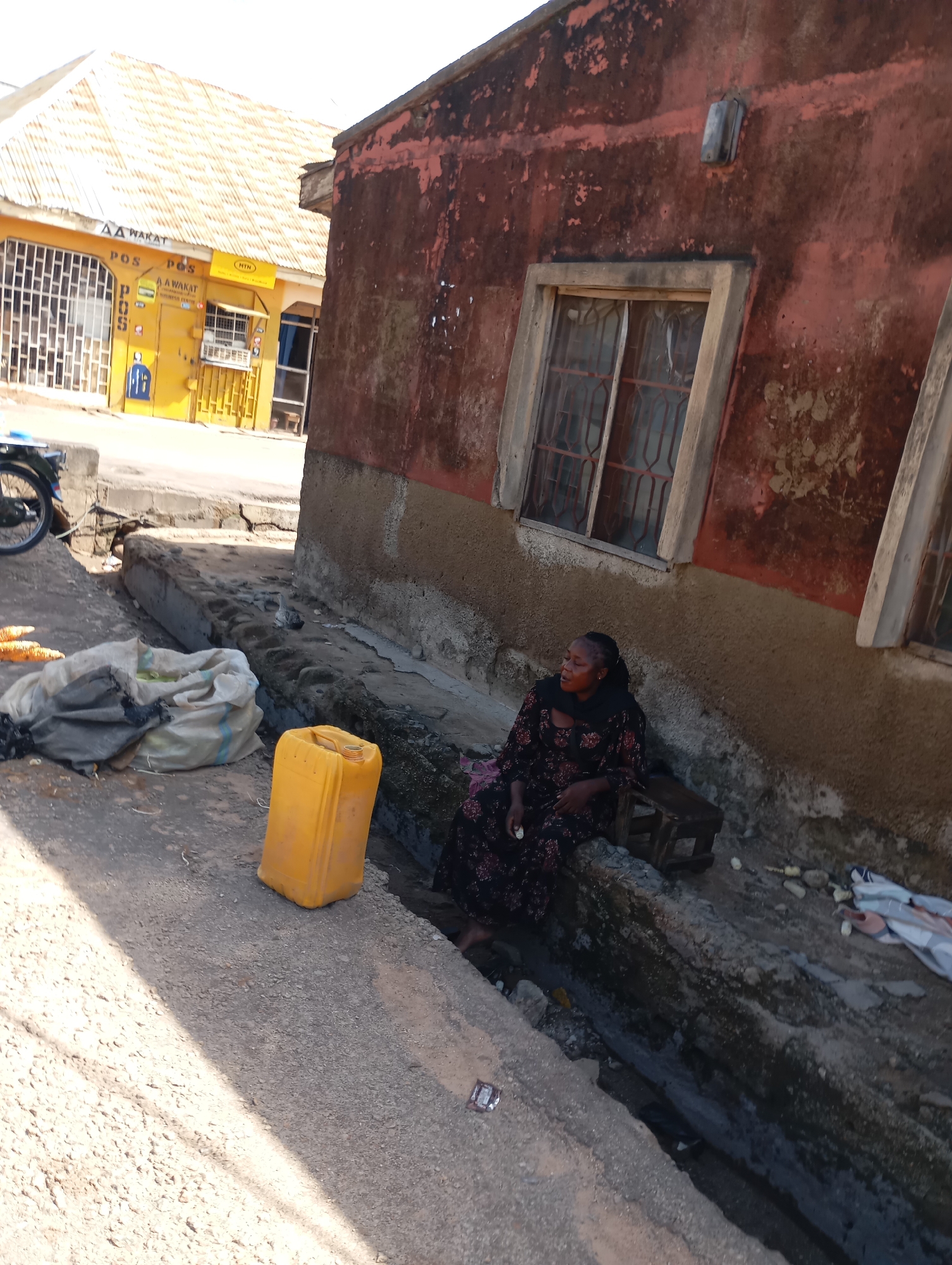 In Shendam, Nigeria, a woman sits on a low wooden stool in a drainage ditch next to a weathered, red-walled building. She is wearing a dark, floral-patterned dress and a black headscarf. Beside her is a yellow plastic container and a large sack. The building has at least two windows with metal grilles and is constructed of concrete and what appears to be plastered mud. Across the street, a yellow building with a corrugated metal roof houses businesses, including one labeled "POS" and another with an "MTN" logo. A portion of a motorcycle is visible on the left side of the image. The scene depicts an outdoor environment with a paved area and the drainage system.