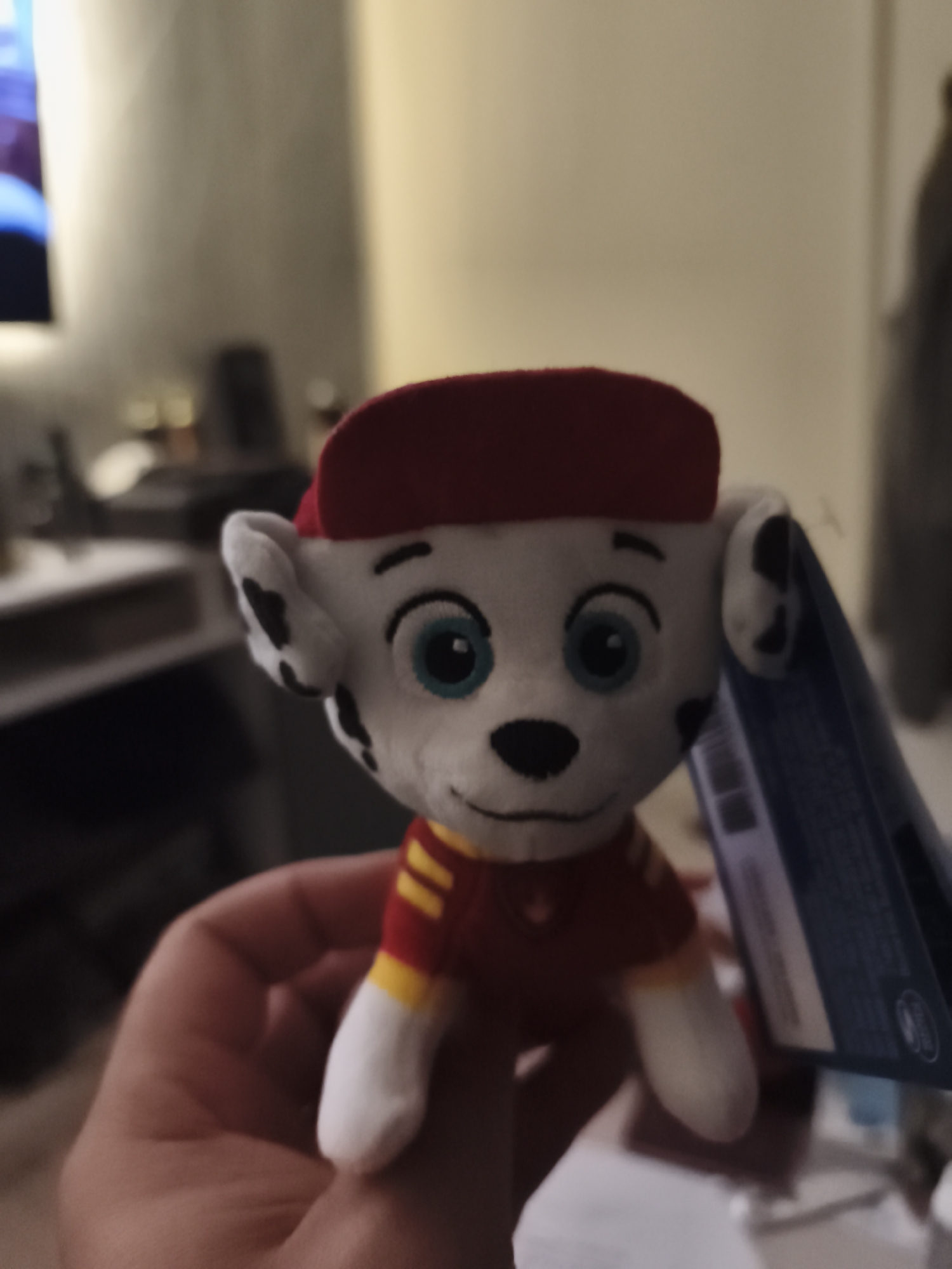 The image features a small plush toy of Marshall from the animated series Paw Patrol, held upright by a person's hand. Marshall is a white Dalmatian puppy wearing a red baseball cap and a red shirt with yellow stripes on the sleeves. He has black spots, bright blue eyes, and a black nose, depicted with a friendly, slightly smiling expression.
The hand, showing parts of the palm and fingers, holds the toy near its midsection. The background is softly blurred, indicating a shallow depth of field. On the left, a bright, out-of-focus blue light source, possibly a television screen, is visible. Other indistinct shapes and ambient lights in the background suggest an indoor setting, likely a domestic living area or bedroom. The overall lighting is dim, implying it might be evening or night. A dark-colored product tag, with small, illegible text and symbols, is attached to the right side of the toy. The scene takes place indoors, within a home in Huntingdonshire, United Kingdom.
