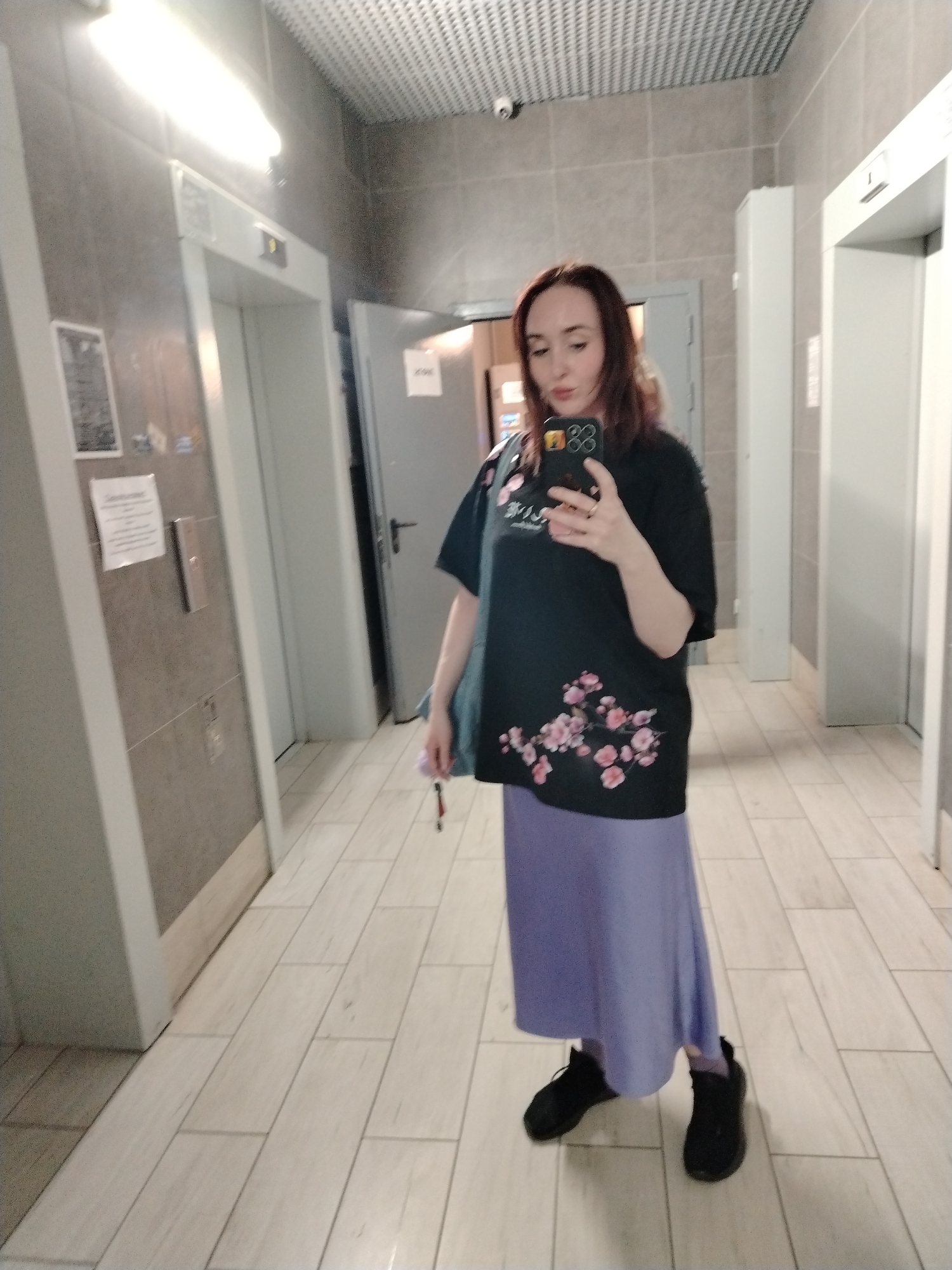 A woman is standing in what appears to be a hallway of an apartment building, taking a selfie with her phone. She is wearing a black t-shirt with pink flower designs, a light purple long skirt, and black sneakers. The hallway has light-colored tiled floors and walls. There are elevators on either side of her, and an open doorway in the background. A small security camera is visible on the ceiling. The photo may have been taken in Izhevsk, Russia, but there are no explicit indicators.