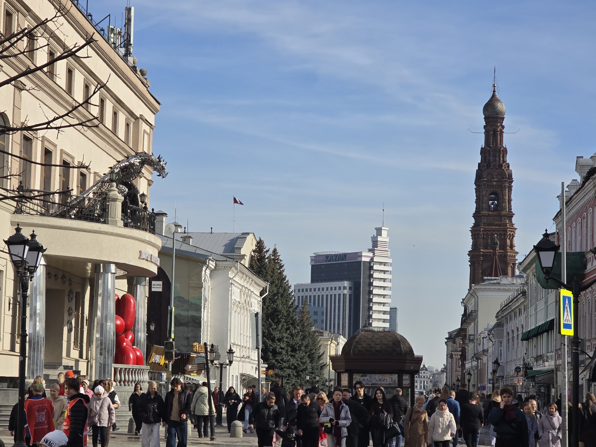 The image depicts a bustling daytime scene on Bauman Street in Kazan, Russia. Numerous pedestrians, dressed in cool-weather jackets and coats, occupy a wide, paved pedestrian thoroughfare.
On the left side of the frame, a multi-story building with a light-colored, classical-style facade features a large, metallic dragon sculpture mounted on a second-floor balcony. Below this, at street level, a cluster of prominent red, bulbous sculptures is visible near an entrance. A black lamppost with two lights stands in the extreme left foreground.
The right side of the street is lined with a row of historic buildings, some featuring ground-level storefronts and awnings. A second black lamppost with a yellow pedestrian crossing sign is visible. Dominating the midground skyline on the right is the ornate, reddish-brown Epiphany Church Bell Tower, distinguished by its multi-tiered structure, golden dome, and spire.
In the center-background, a modern high-rise building with "GRAND KAZAN HOTEL" visible on its facade stands prominently. Several dark green evergreen trees are interspersed among the buildings further down the street. A small Russian flag flies from a distant pole near the center of the sky. The sky is largely clear blue with some faint clouds.