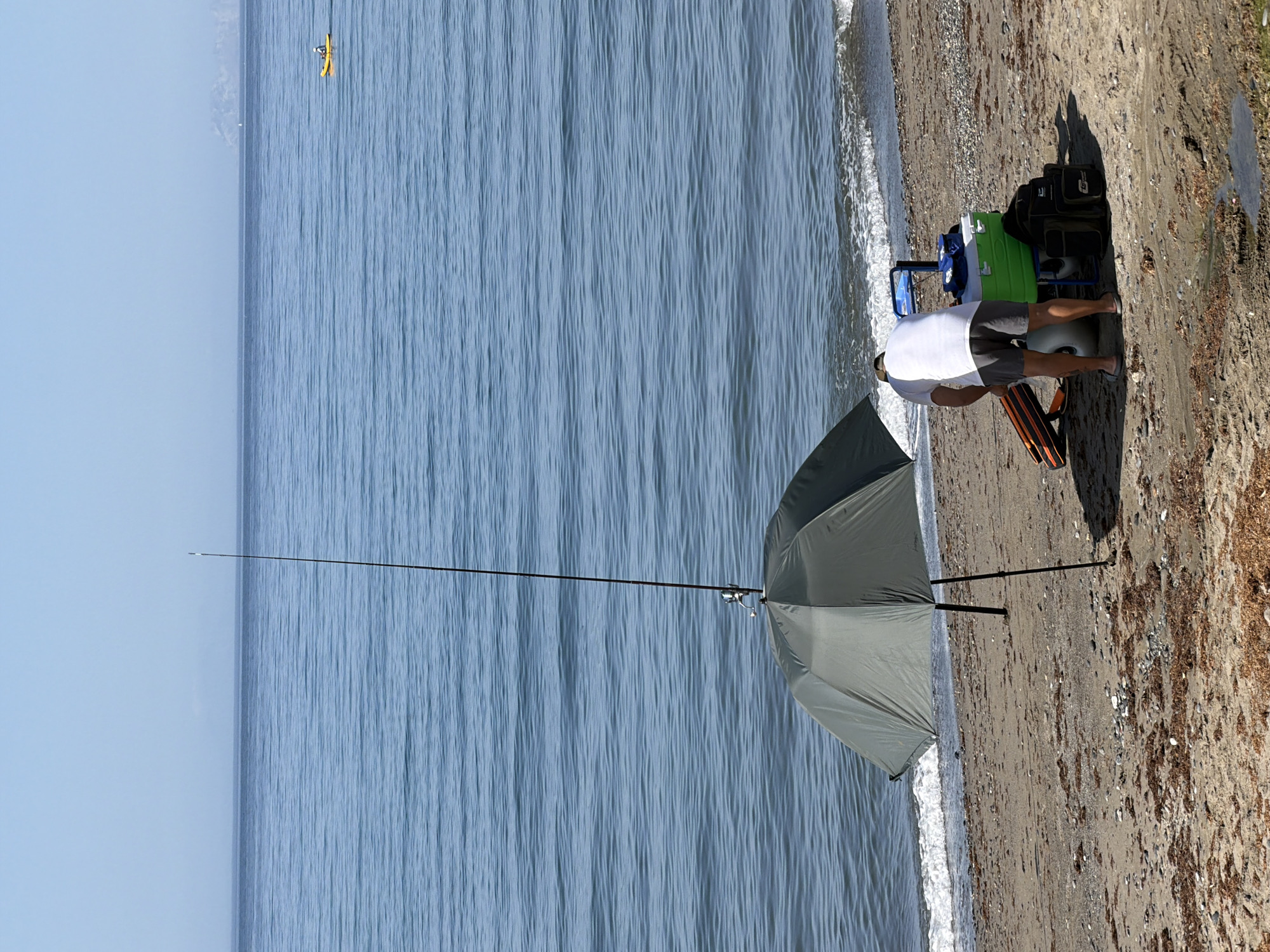 A man is bent over a fishing trolley on a beach in Estepona, Spain. He is wearing a white t-shirt and grey shorts. Next to him is a green cooler and several fishing bags. An open grey umbrella and a fishing rod stand upright on the sand behind him. The ocean is calm and blue, with a few small waves reaching the shore. In the distance, a yellow kayak with a person in it is visible on the water, and a hazy landmass can be seen on the horizon. The beach is covered in sand and small pebbles, with scattered seaweed.