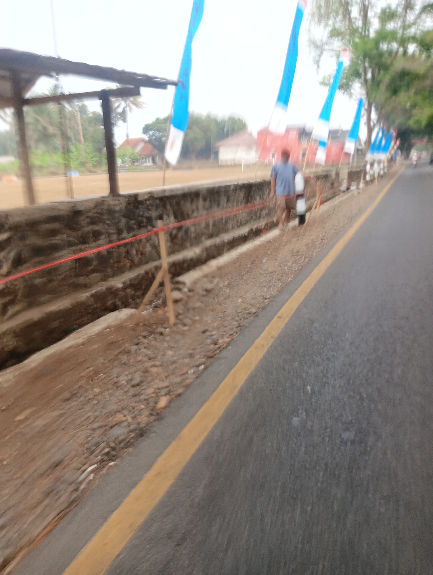 The image depicts a street scene in Manggari, Indonesia, with several blue and white flags lining the roadside. A person, wearing a blue t-shirt and brown shorts, stands near the flags, interacting with a white object. The street is paved and has a yellow line marking the lane. To the left of the road, there is a retaining wall and some construction or landscaping work in progress, with visible wooden stakes and red tape. Beyond the wall, there is an open field, some houses with red roofs, and trees. The sky is overcast. The image is blurred, suggesting movement during capture.