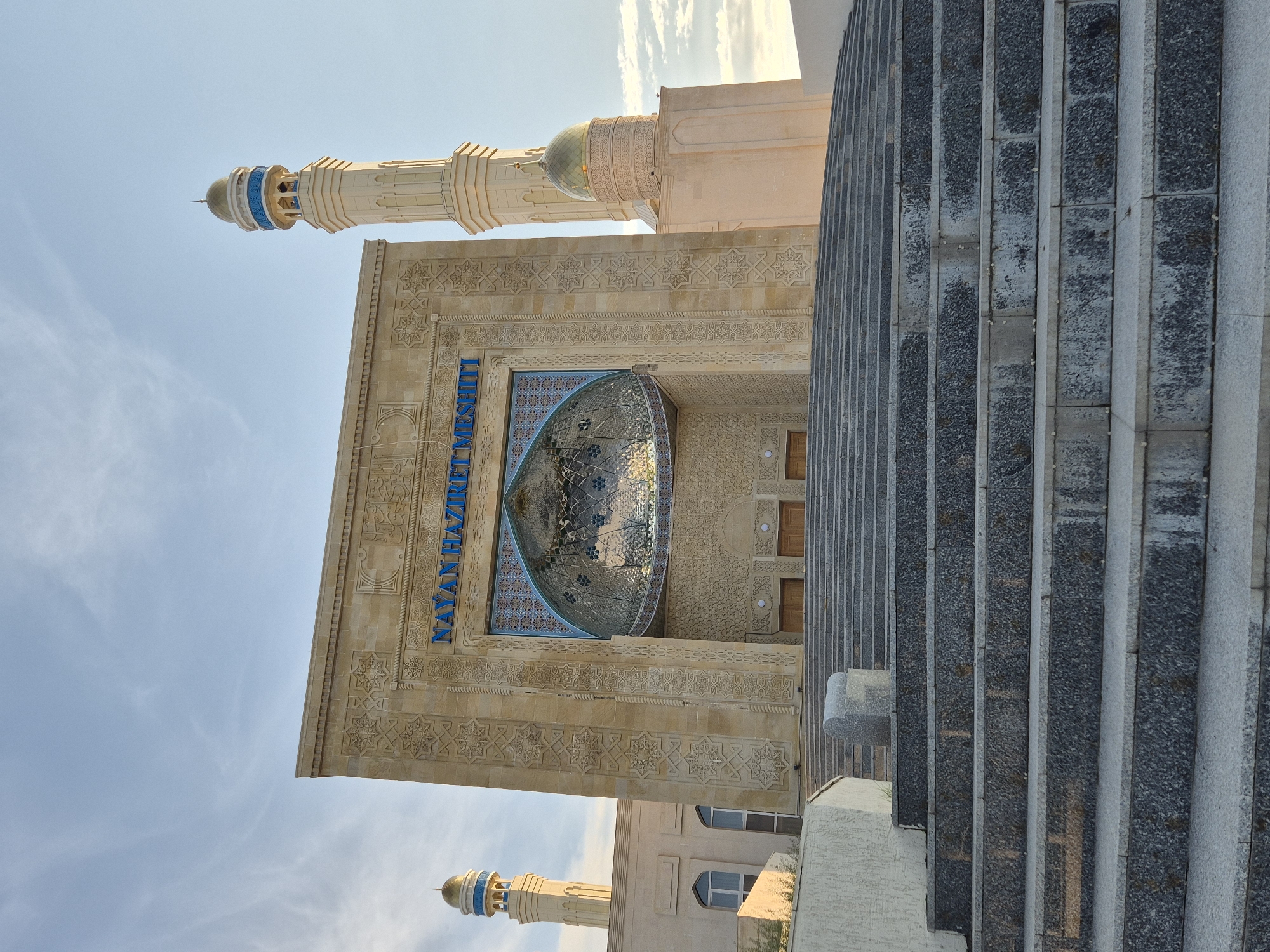 The image shows the exterior of the Nayan Hazrat Mosque in Kokshetau, Kazakhstan. The mosque features a prominent facade with intricate geometric patterns and a large, mirrored, domed entrance. A minaret with a blue and gold decorative top is visible. A set of stairs made of dark gray stone is on the right side of the image. The sky is visible with some clouds.