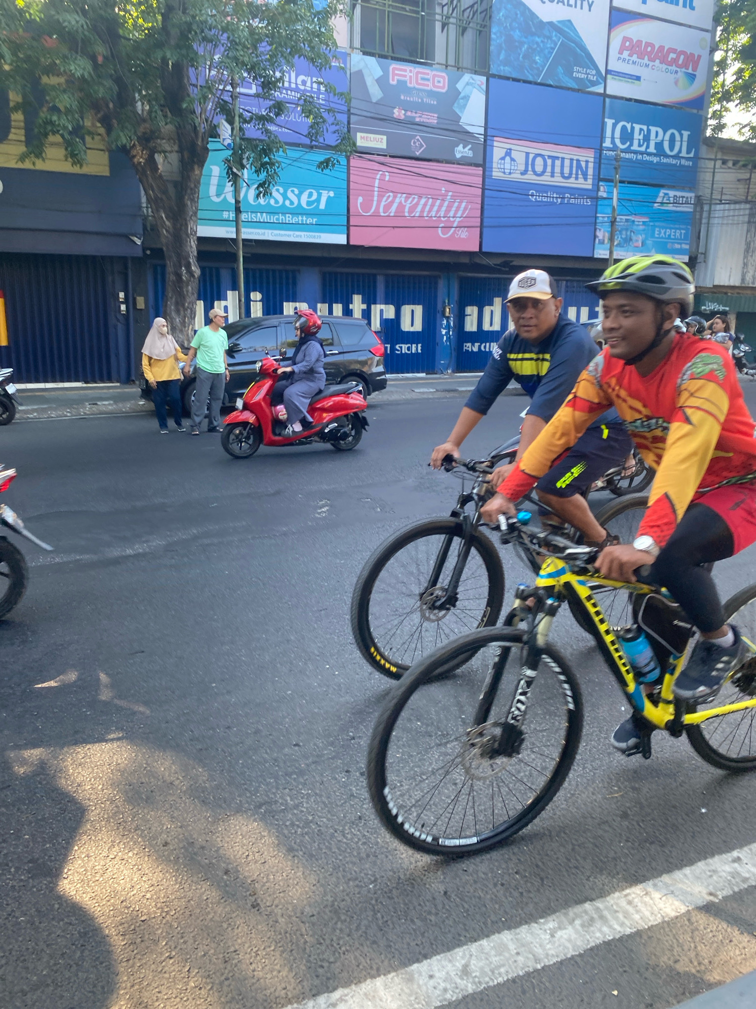 The image captures a street scene in Kediri City, Indonesia, characterized by a bustling atmosphere with several vehicles and pedestrians. The foreground features two individuals riding bicycles. The person on the right, wearing a yellow and red long-sleeved jersey, a helmet, and black leggings, is actively cycling. To their left, another person, dressed in a blue long-sleeved shirt, dark shorts, and a white cap, is also on a bicycle. In the middle ground, a red scooter is stopped, with a person wearing a blue shirt and a helmet seated on it. Beside the scooter, two pedestrians, one in a yellow hijab and the other in a light green t-shirt and grey pants, are standing near a dark-colored car. The background is dominated by a large building with numerous advertisements displayed on its facade, including brands like "PARAGON," "JOTUN," and "ICEPOL." The street is paved and appears wet in places, reflecting the sunlight. Trees line the street, and the overall lighting suggests it is daytime.