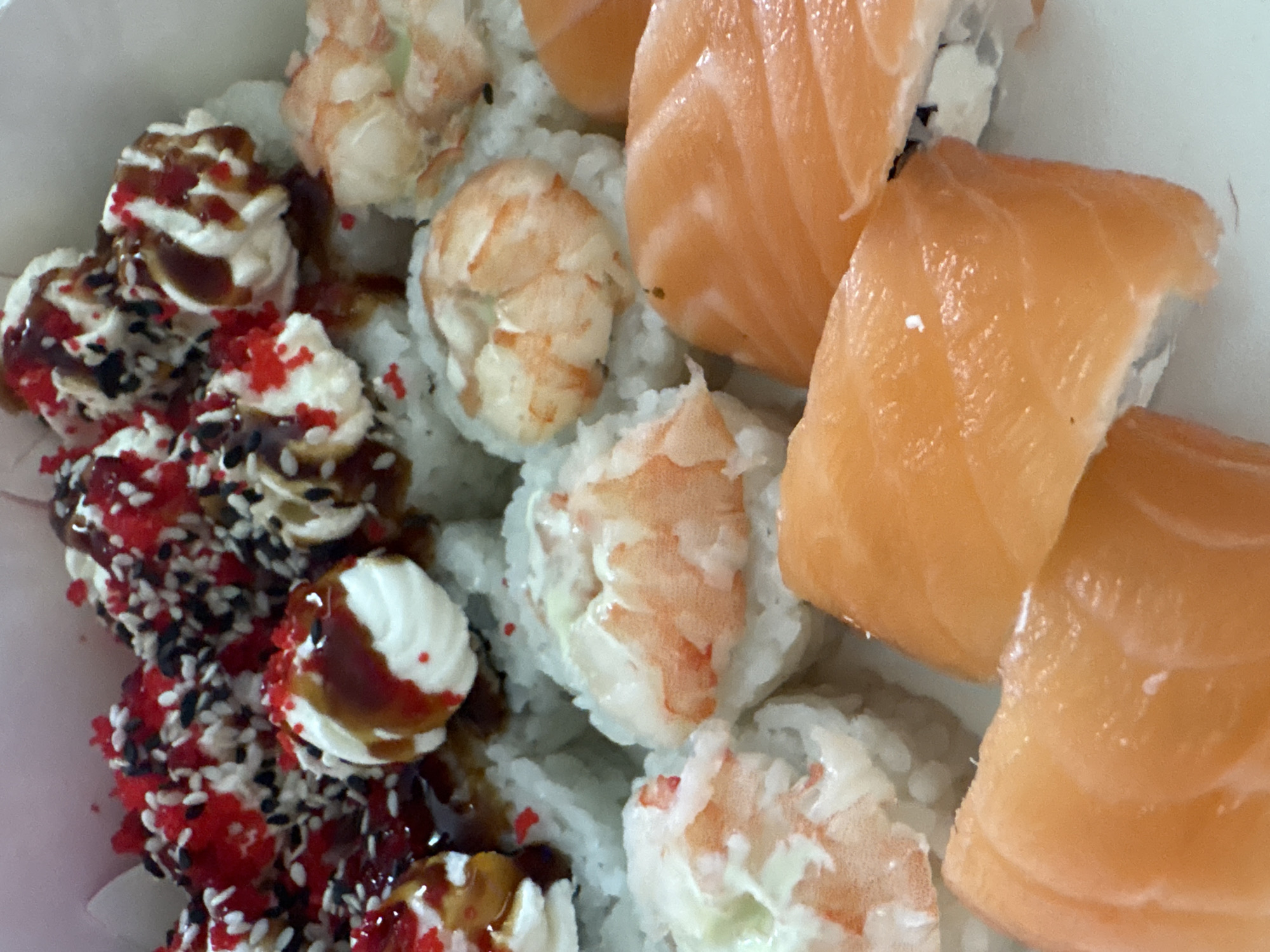 The image shows a close-up view of sushi rolls arranged in a container. On the left, there are rolls topped with cream cheese, a dark sauce, and red and black sprinkles. In the center, there are rolls with shrimp. On the right, there are rolls wrapped in salmon. The background is blurred, suggesting a shallow depth of field. The overall lighting is diffused, providing even illumination across the sushi. There is no visible information to suggest the location is in the city of Chaikovsky, Russia.